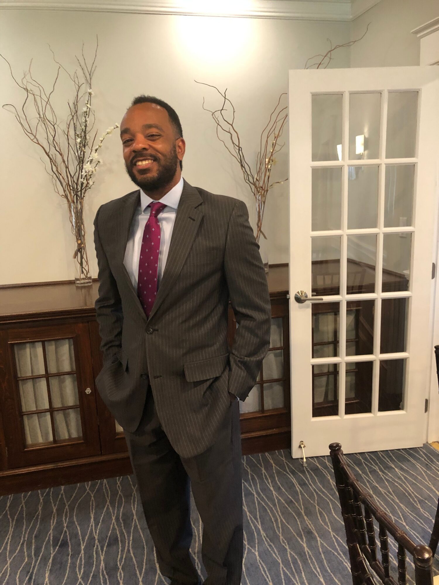 Man in suit standing indoors with decorative branches and glass cabinet behind him.