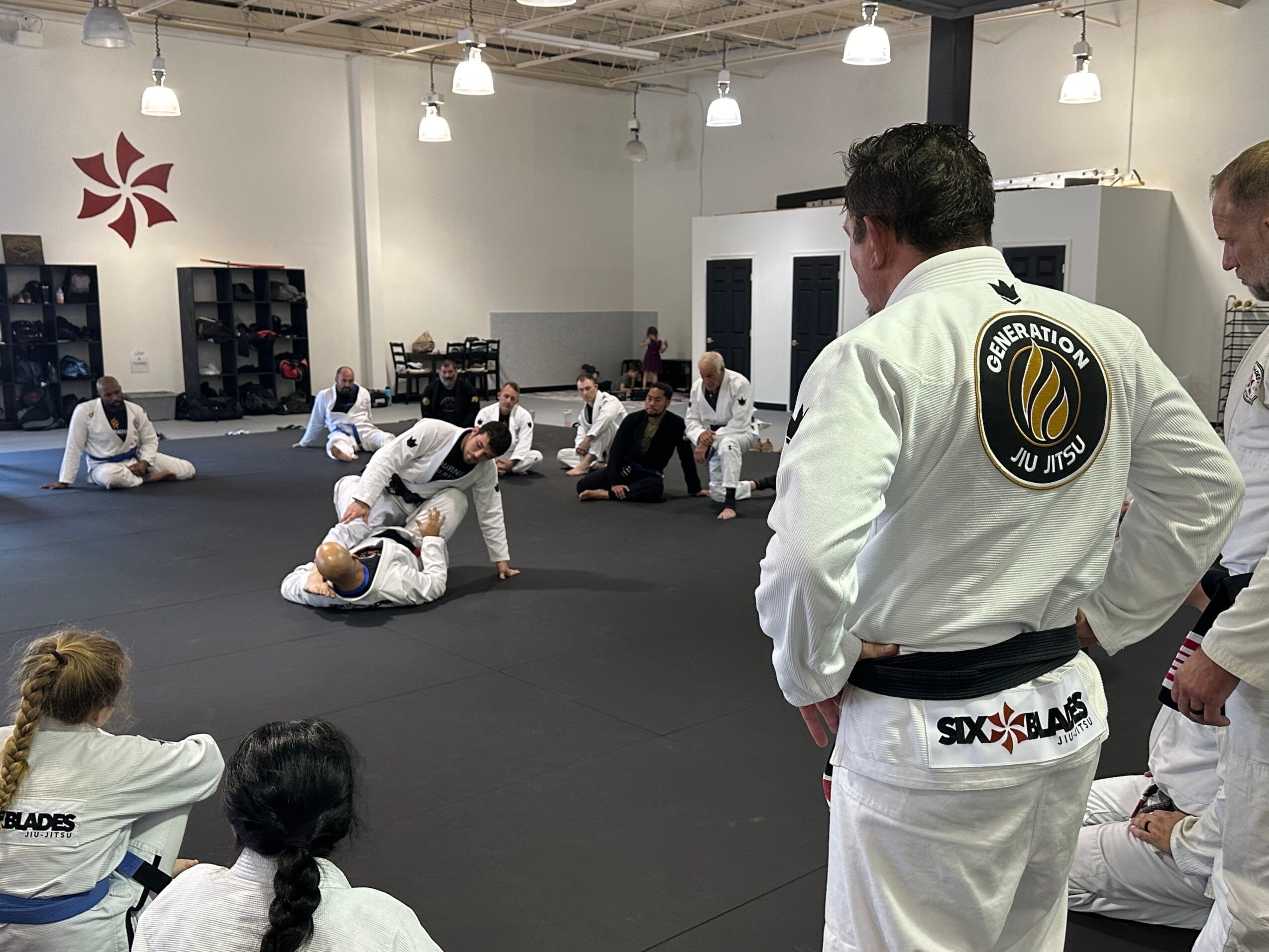 People practicing Brazilian Jiu-Jitsu on mats in a training room with instructors observing.