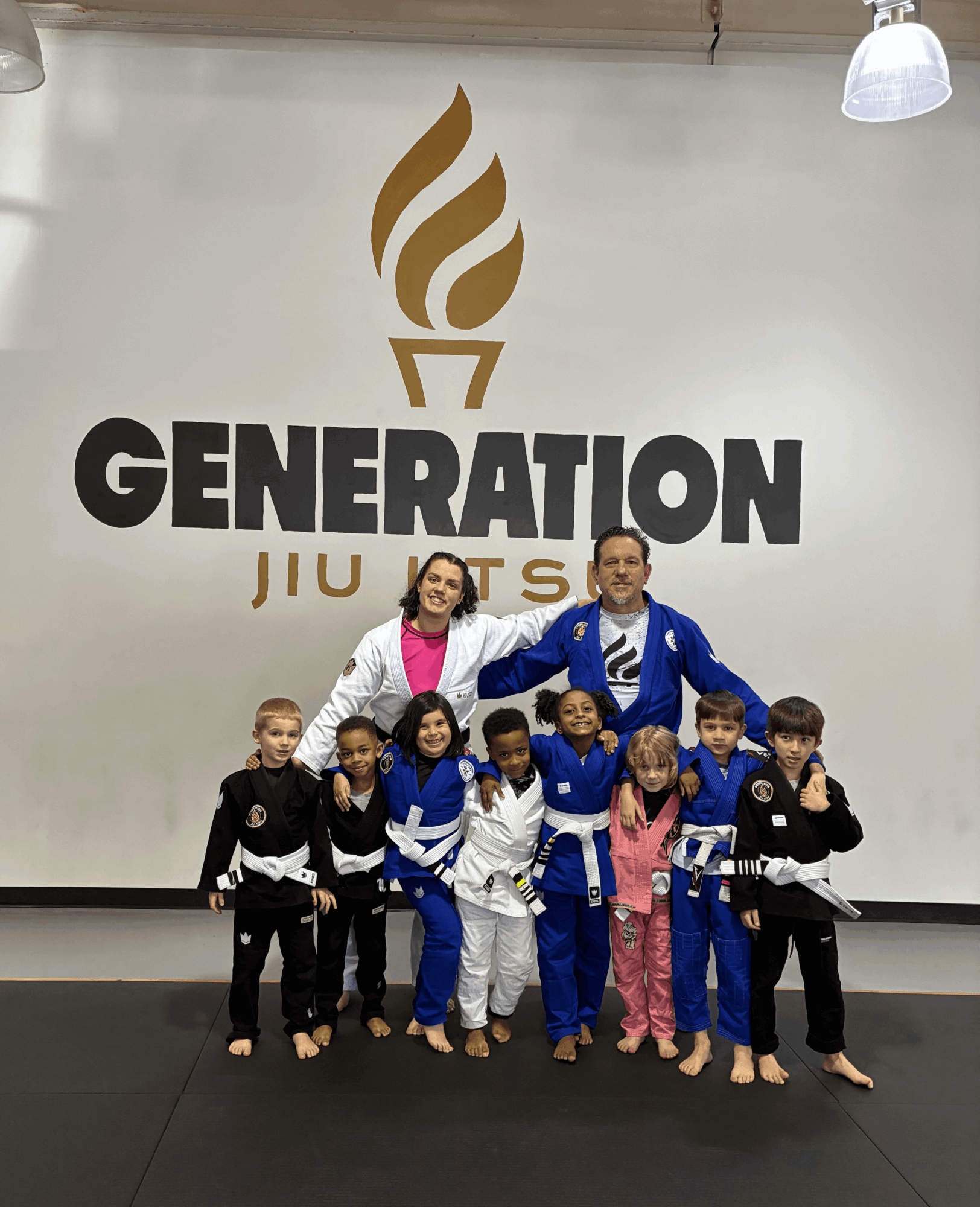 Group of children and two adults in martial arts uniforms standing in front of a wall with a logo and text, smiling.