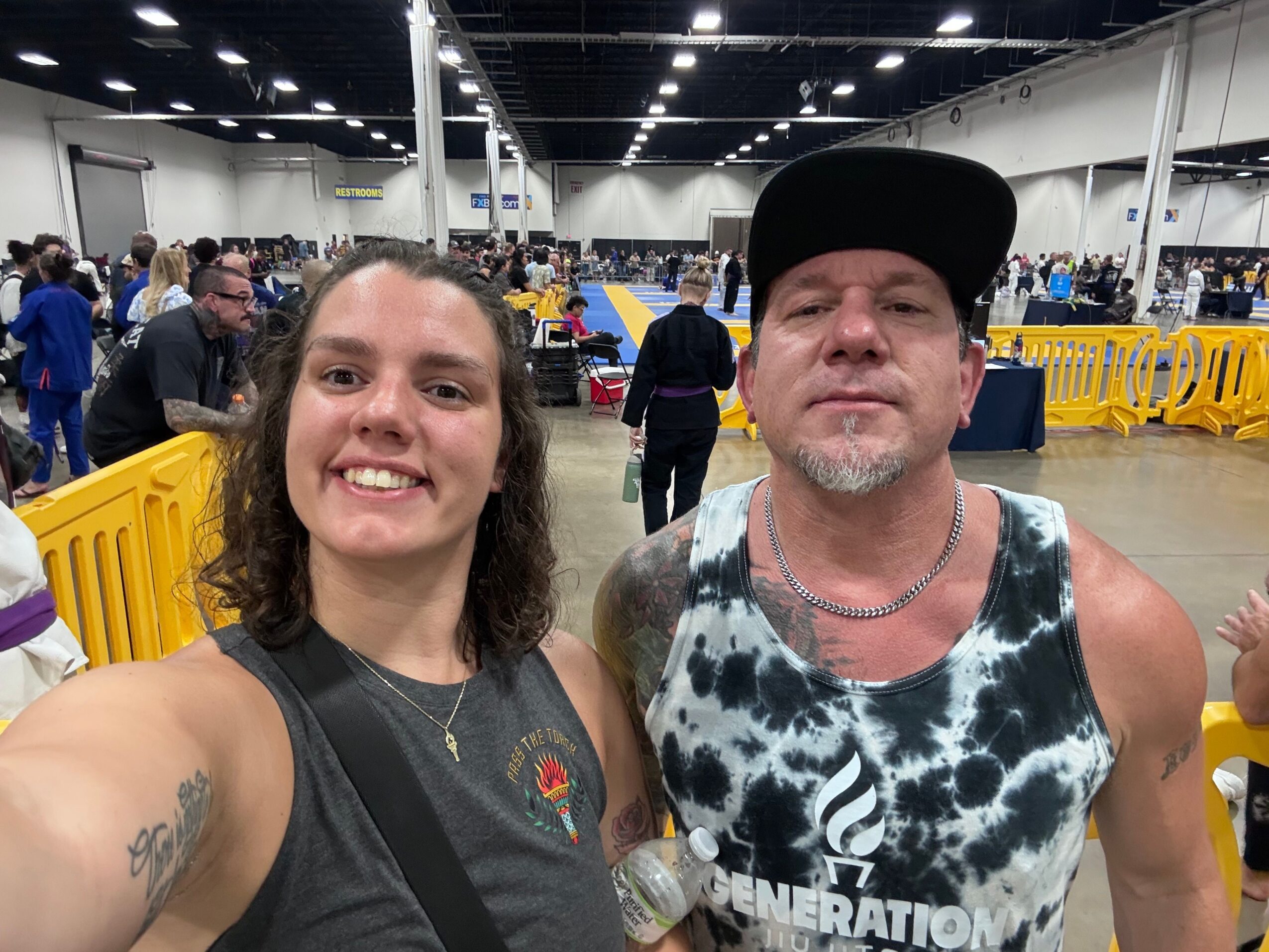 Two people taking a selfie in a crowded indoor space with yellow and blue objects in the background.