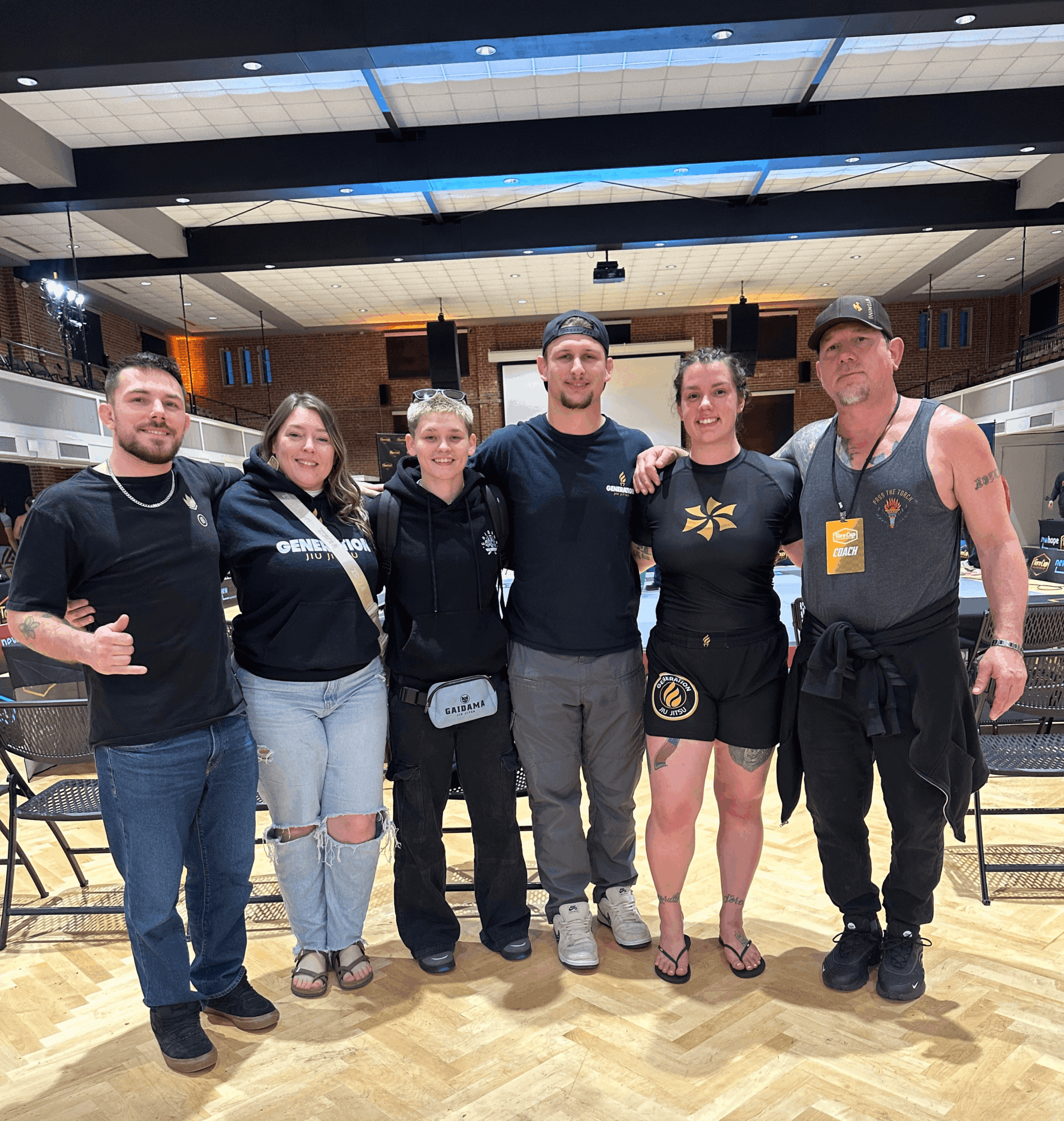 Six people standing together in a room, smiling, with a stage and seating in the background.