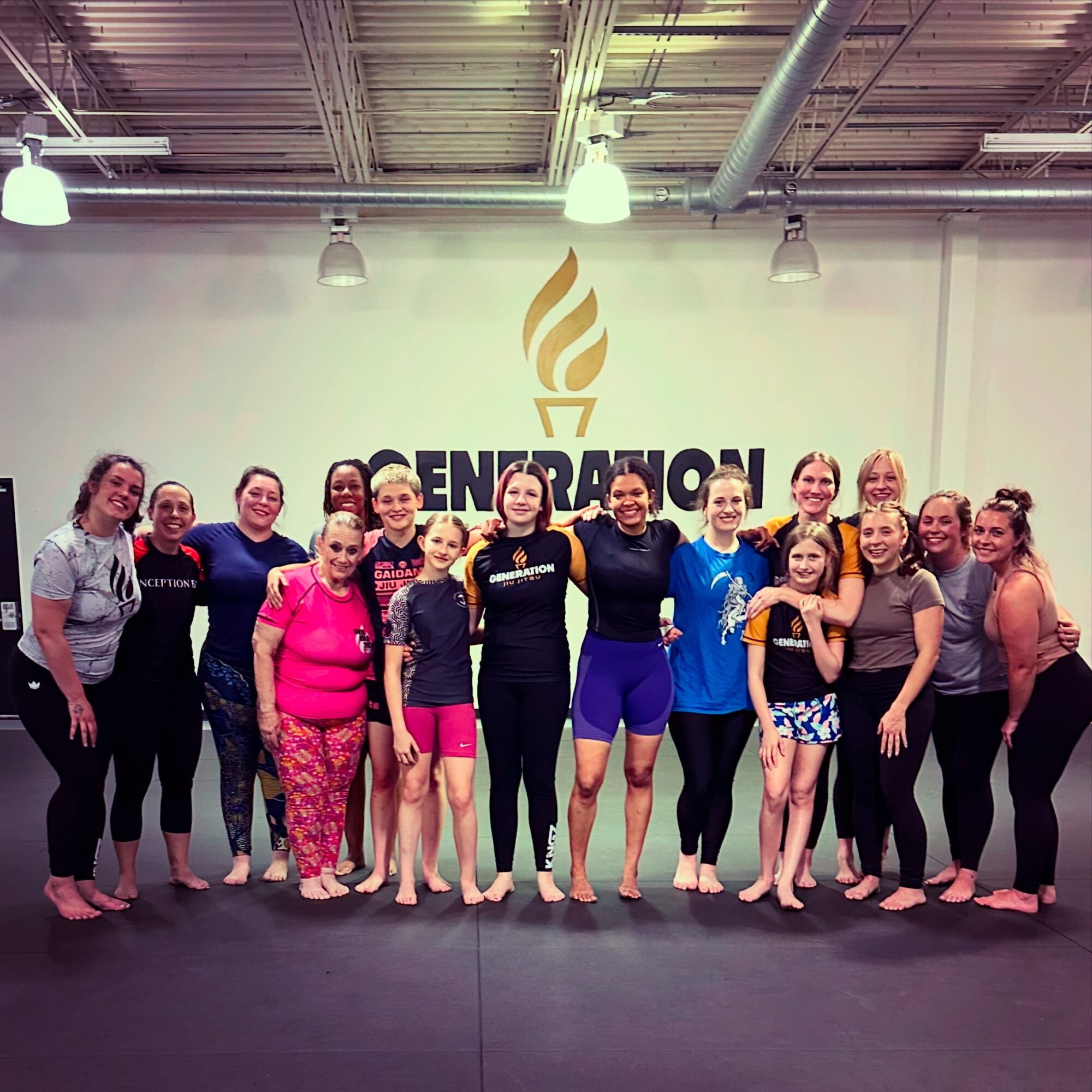 Group of women standing together in a gym, smiling, with a logo and the word 'ENERGIZE' on the wall behind them.