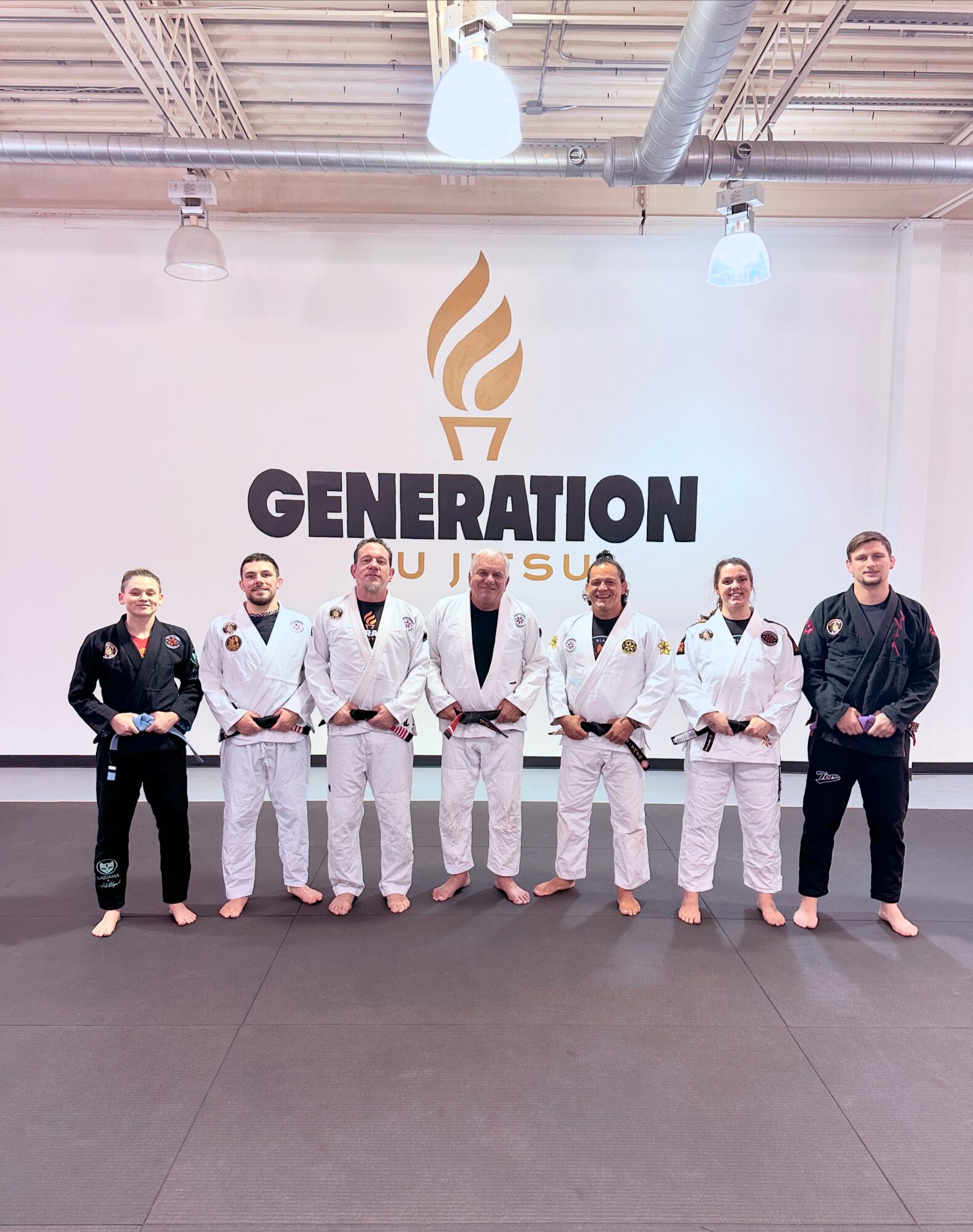 Seven people in martial arts uniforms stand in front of a wall with a logo and the word 'GENERATION'.