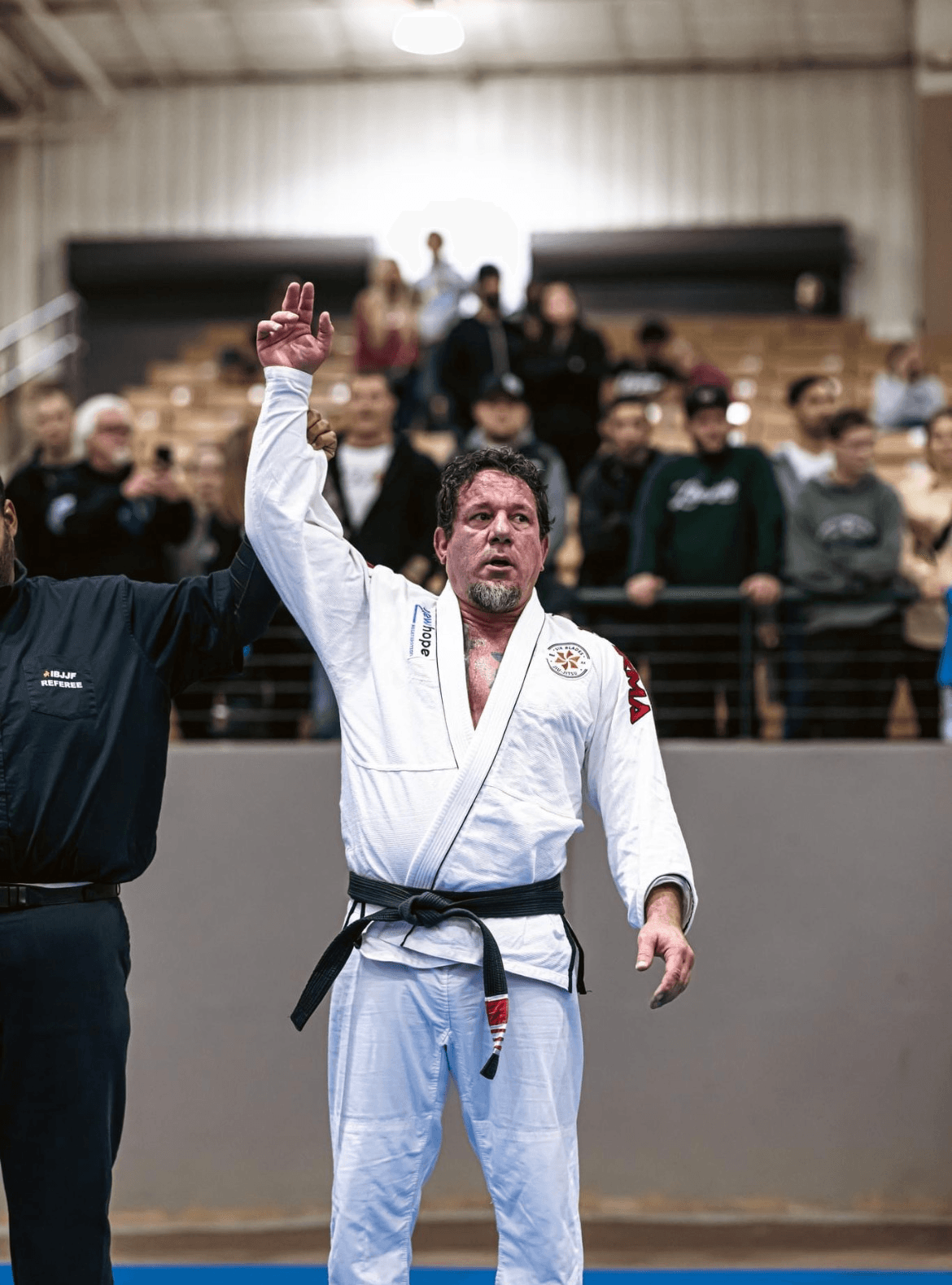 Judo competitor in a white gi with a black belt raising his hand, crowd in the background, indoor sports venue.
