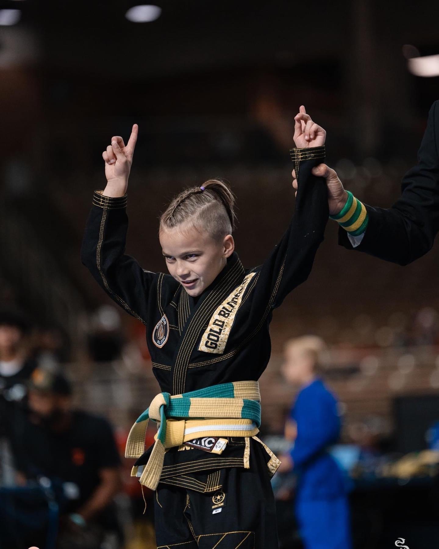 Young girl in martial arts uniform with a gold belt, raising both arms in victory, smiling.
