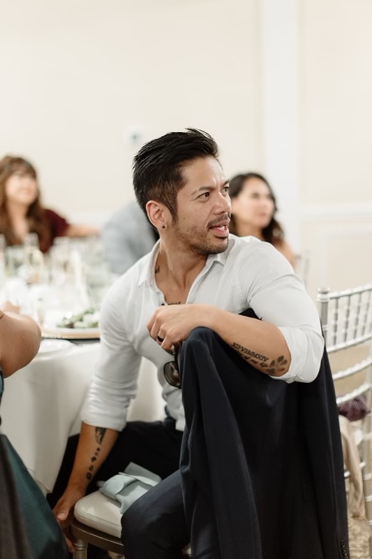 Man with dark hair and beard sitting at a table, smiling, with women in the background, in a bright room.