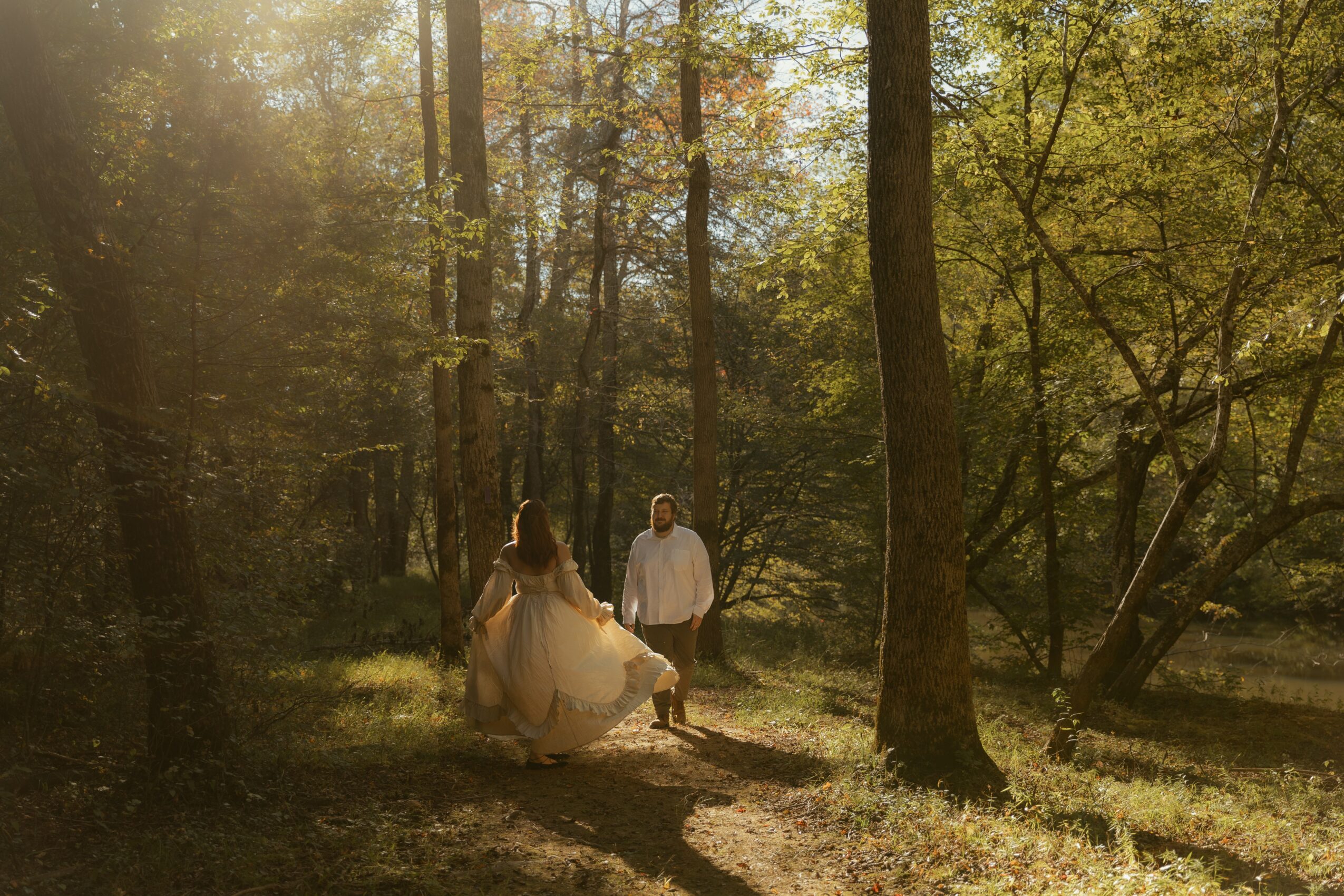Two people walking in a sunlit forest, one in a light dress and the other in casual clothing, surrounded by trees.