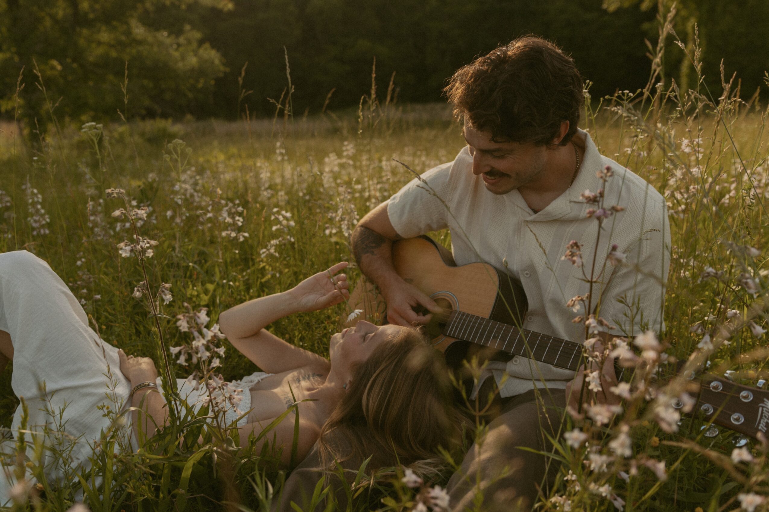 A man plays guitar while a woman lies in grass with flowers, outdoors in a field during daytime.