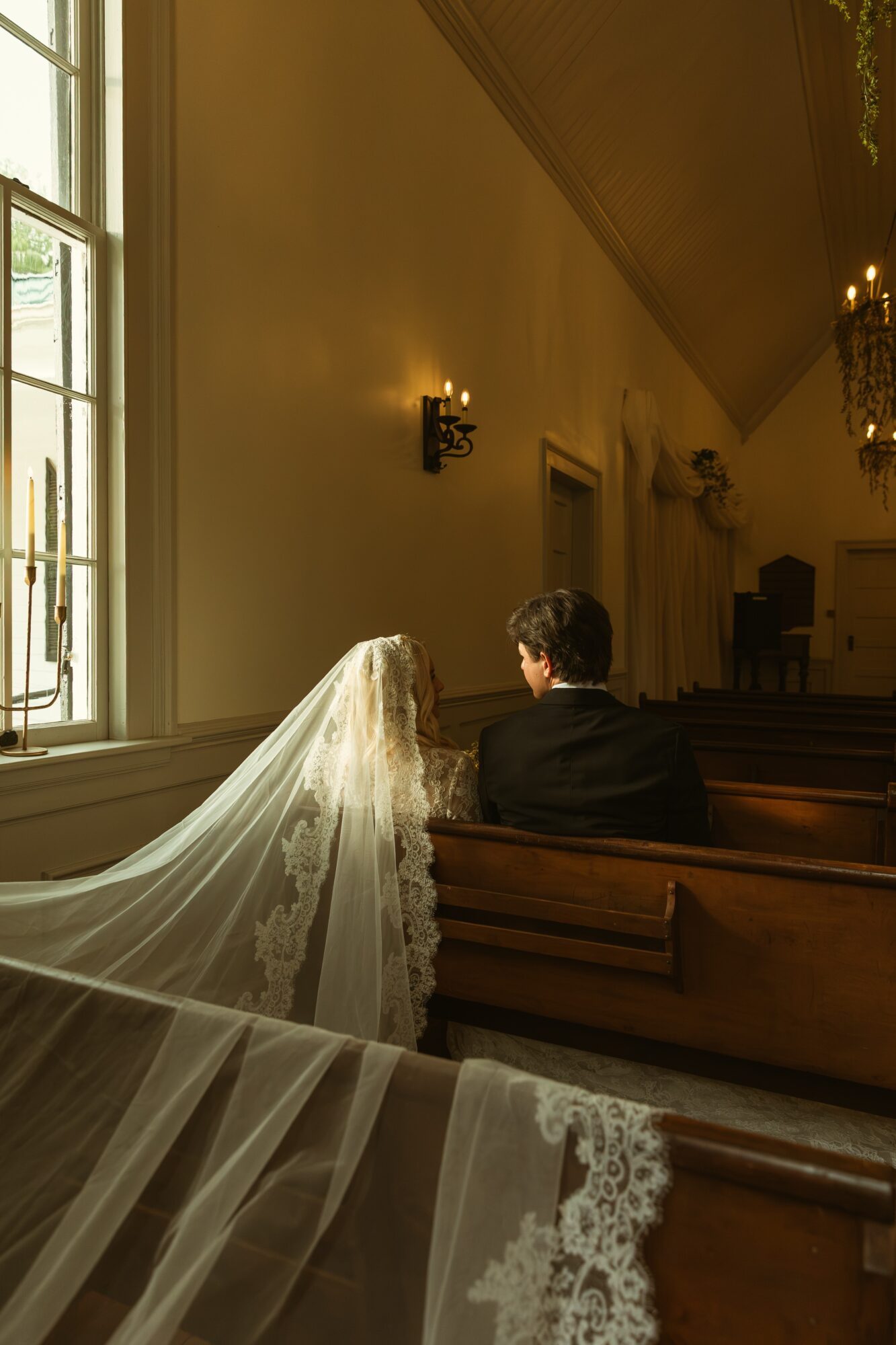 Bride and groom sitting in church pews, bride's veil draped over pew, interior with large window and wall sconces