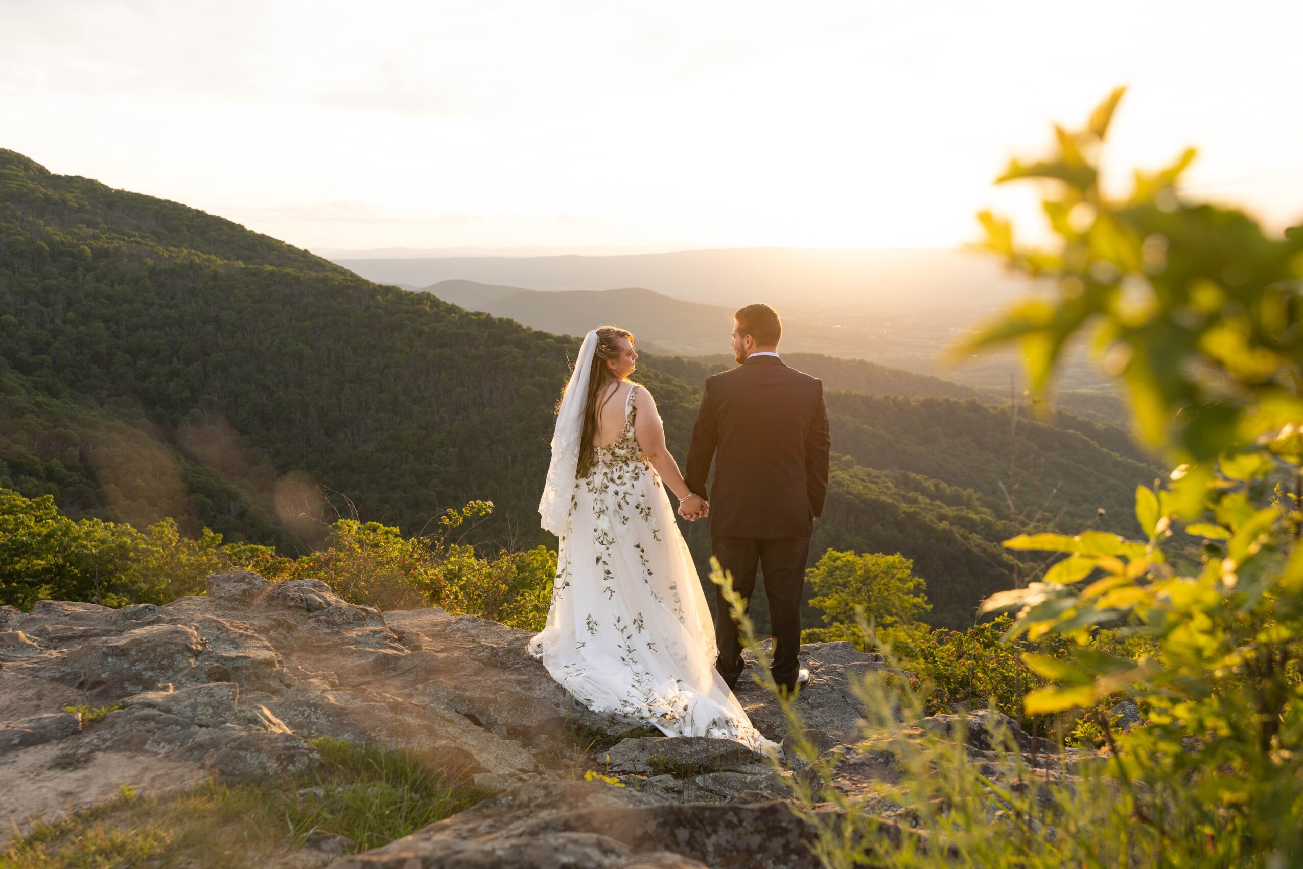 A bride and groom walk together on a rocky hilltop with a scenic landscape and sunset in the background.