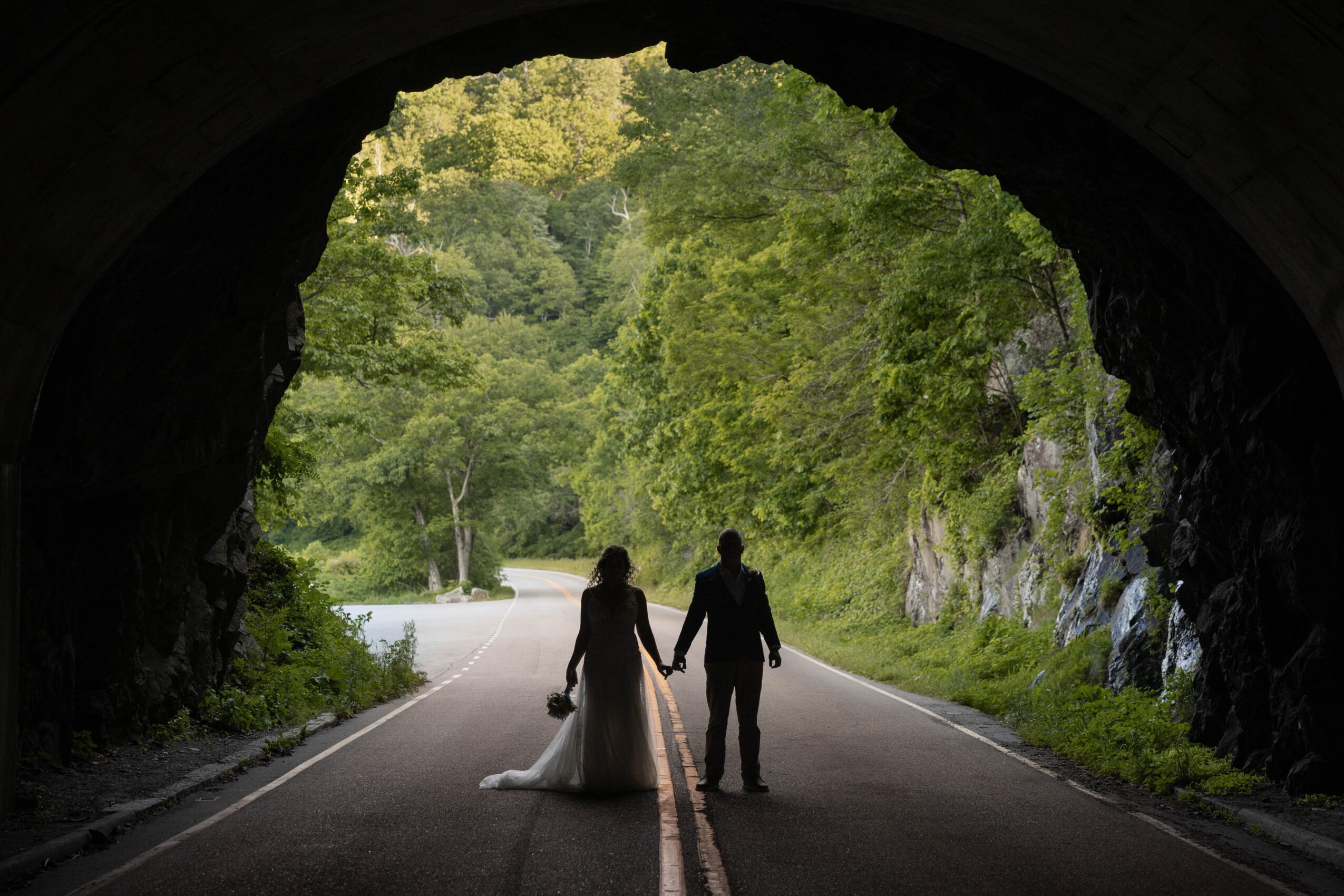 Silhouette of a couple holding hands walking through a tunnel onto a road surrounded by greenery.