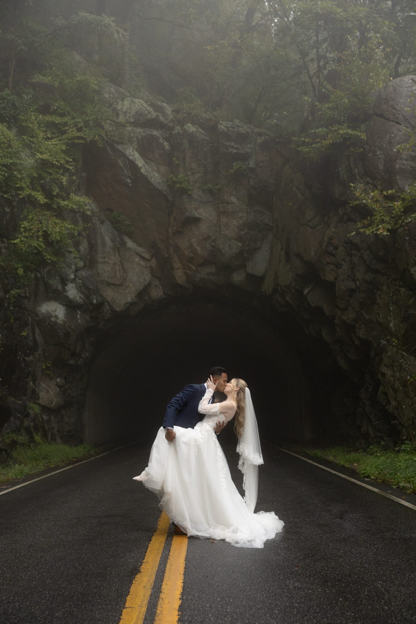 A bride and groom kiss in the middle of a road under a rocky tunnel entrance surrounded by trees.