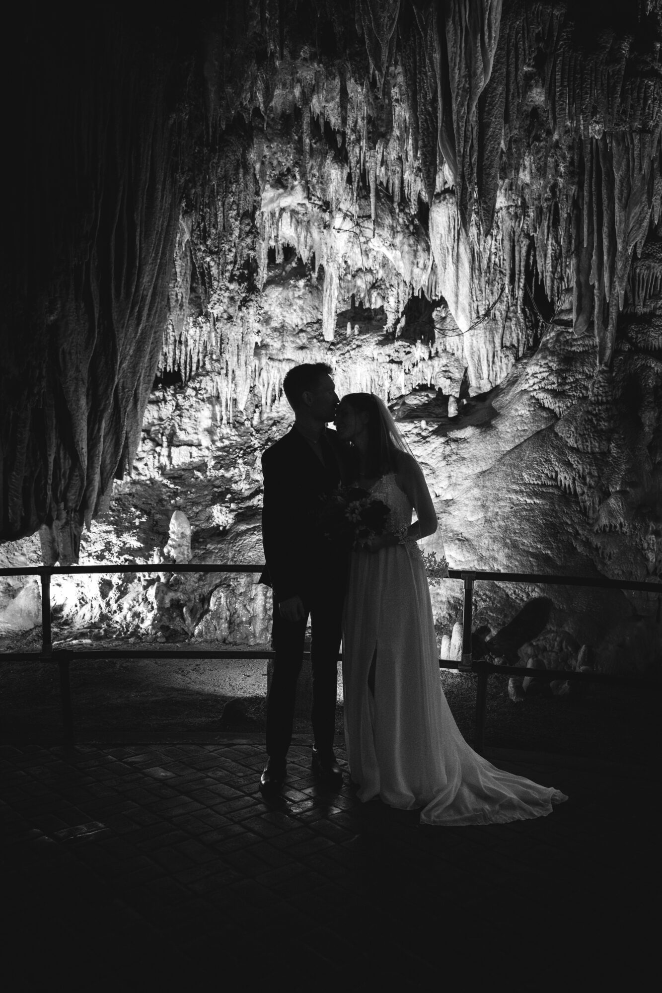 Silhouettes of a couple standing in front of a cave with stalactites and stalagmites, illuminated from behind, in black and white.