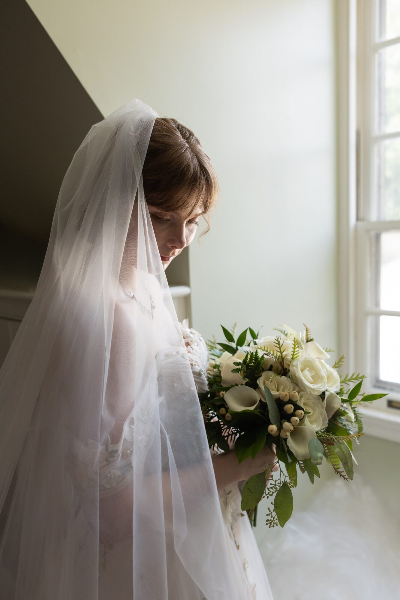 Bride with veil holding a bouquet of white roses and greenery, standing near a window with sunlight, looking down.
