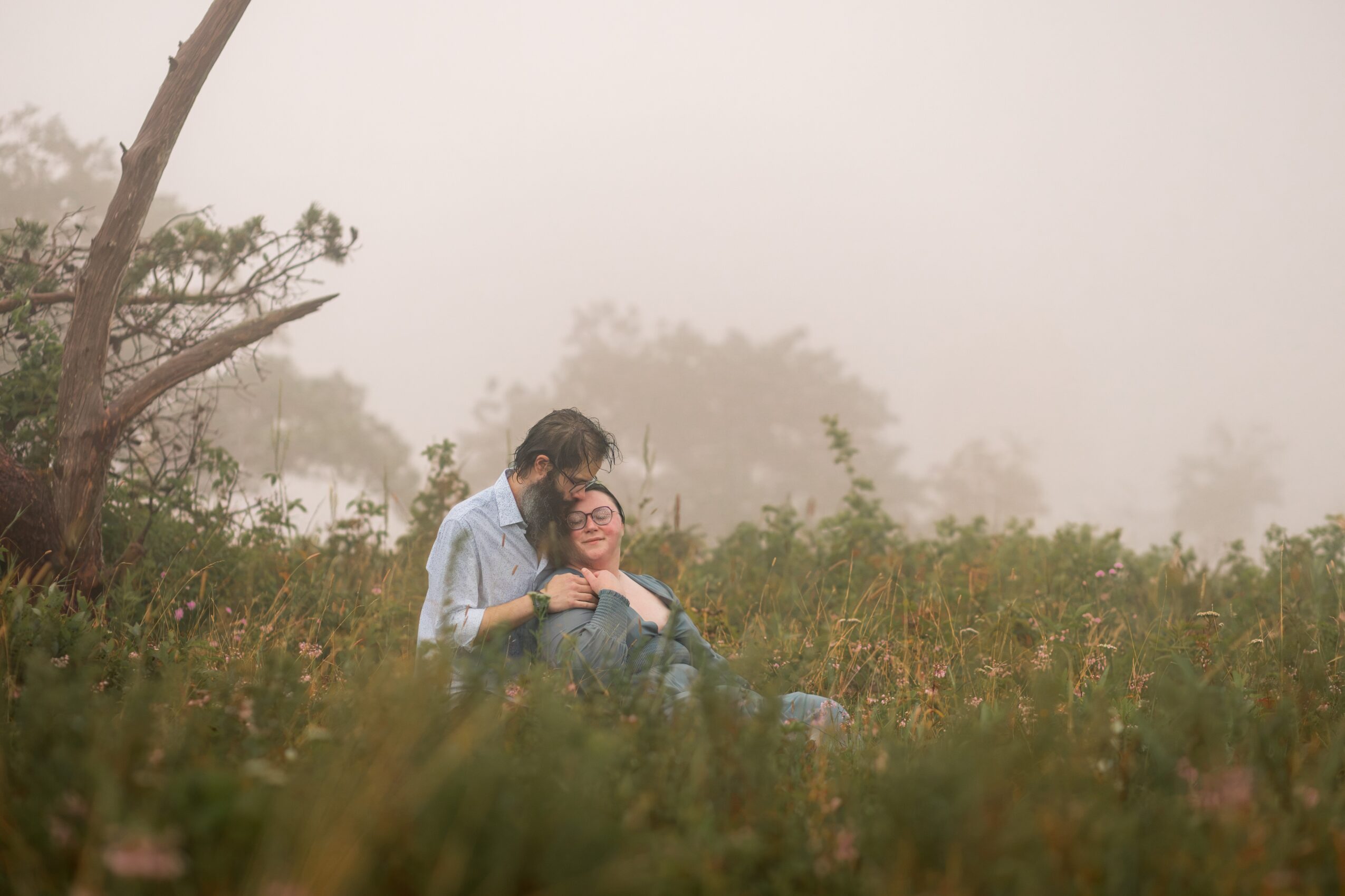 Two people sitting in a grassy field with foggy background, one person leaning on the other's shoulder, trees in distance.