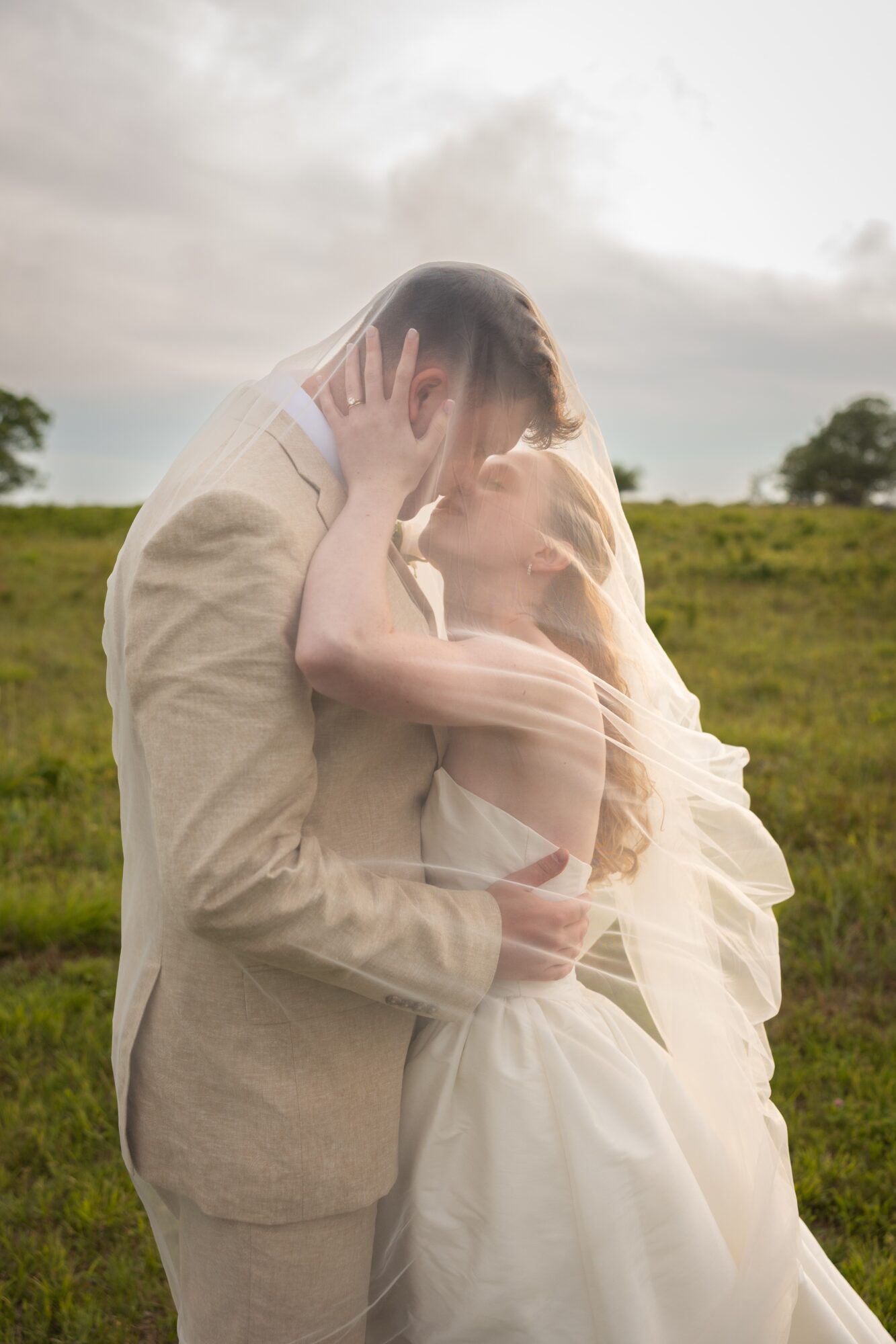 A couple in wedding attire embracing outdoors with a cloudy sky and green field background.
