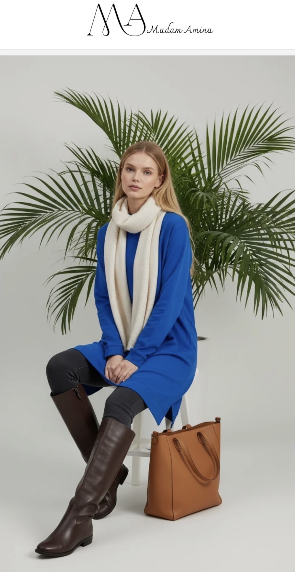 Woman sitting with legs crossed, wearing a blue coat, white scarf, and brown boots, with a large plant behind her.
