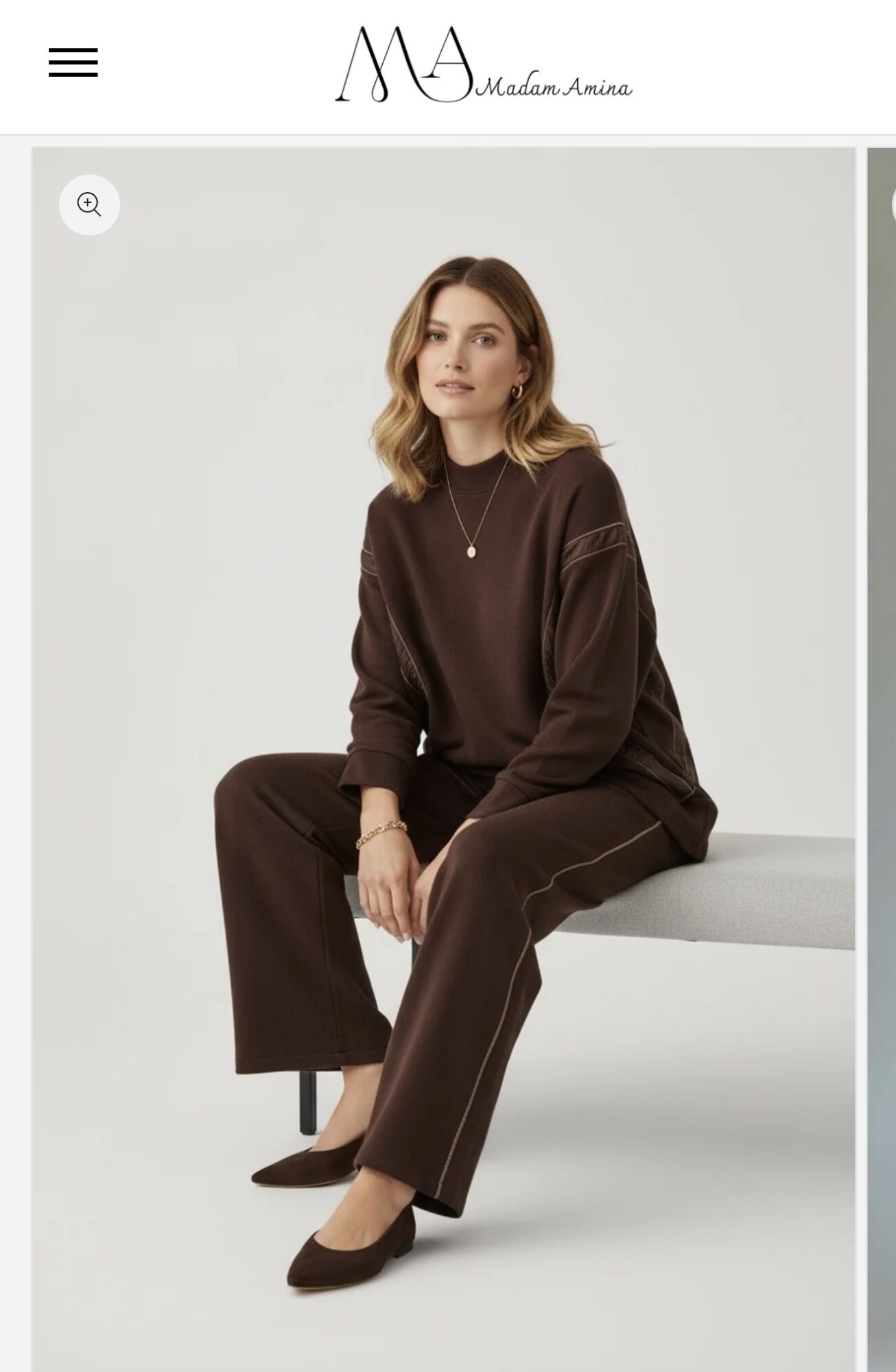 Woman sitting on a bench wearing a dark brown outfit with a necklace, neutral background, relaxed pose.