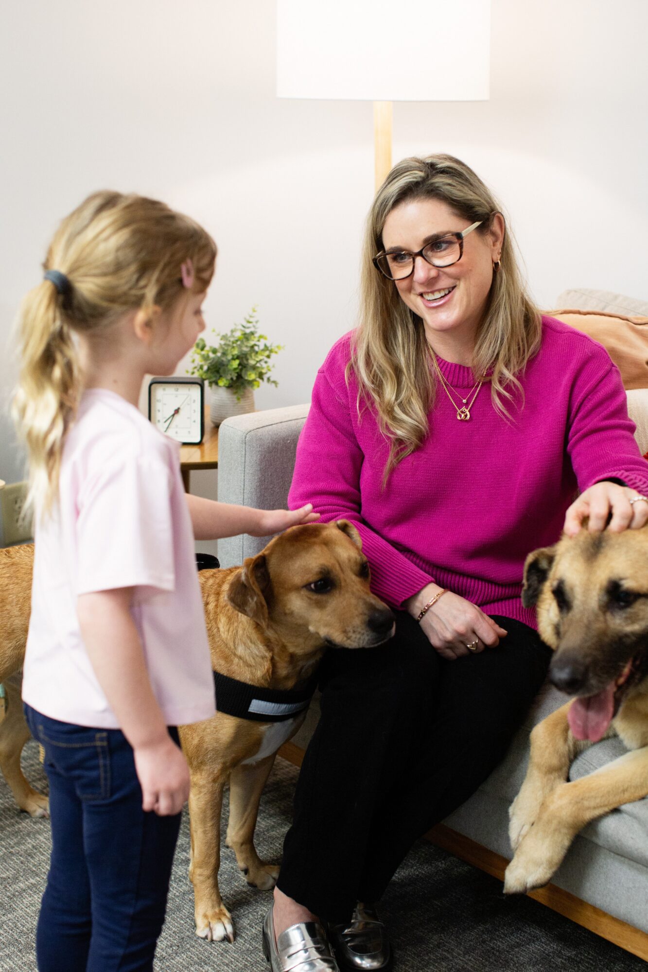 Woman with glasses smiling at girl, two dogs sitting on her lap, in a cozy living room with a lamp and plant.