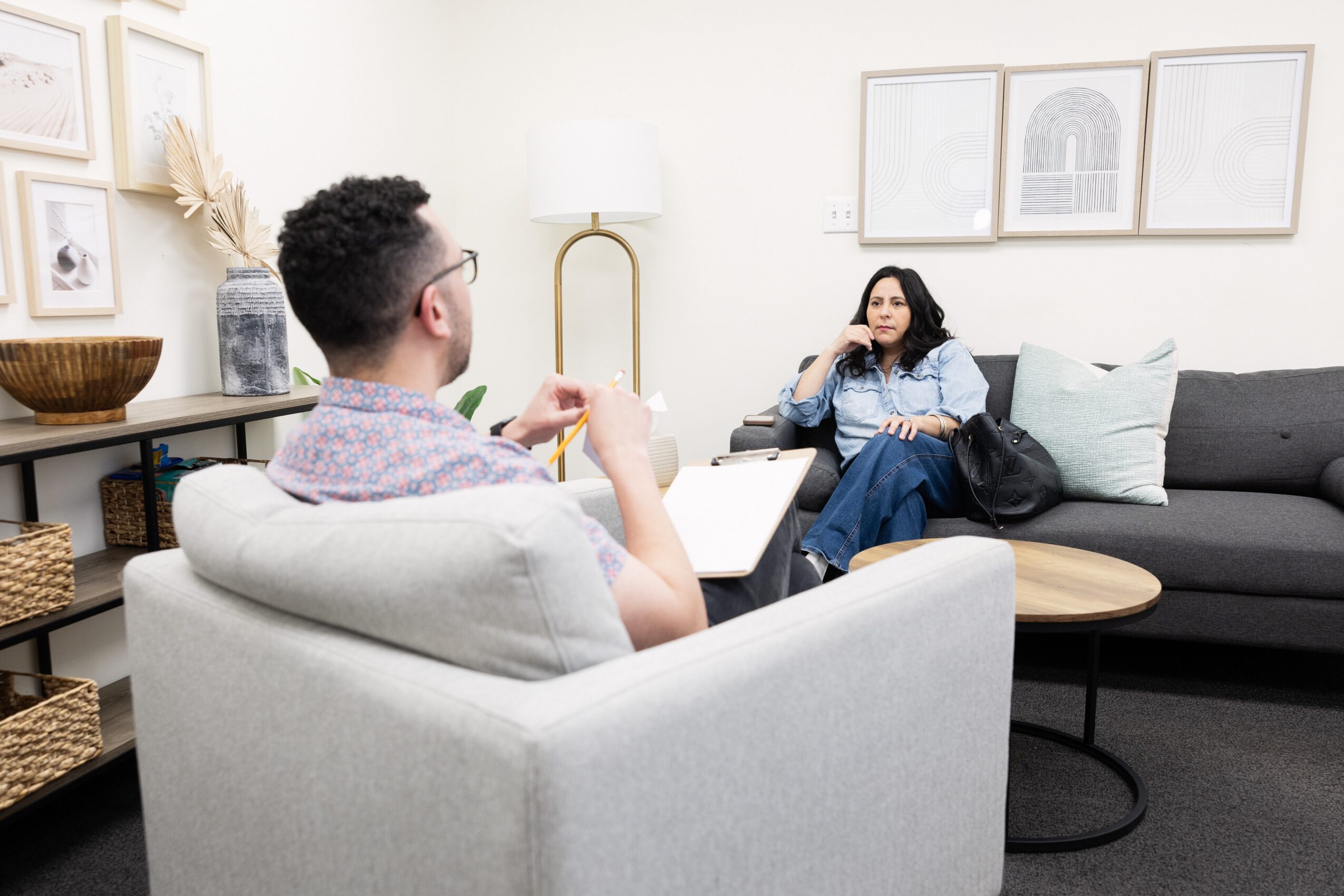 Two people sitting in a living room, one on a white armchair and the other on a dark sofa, talking.