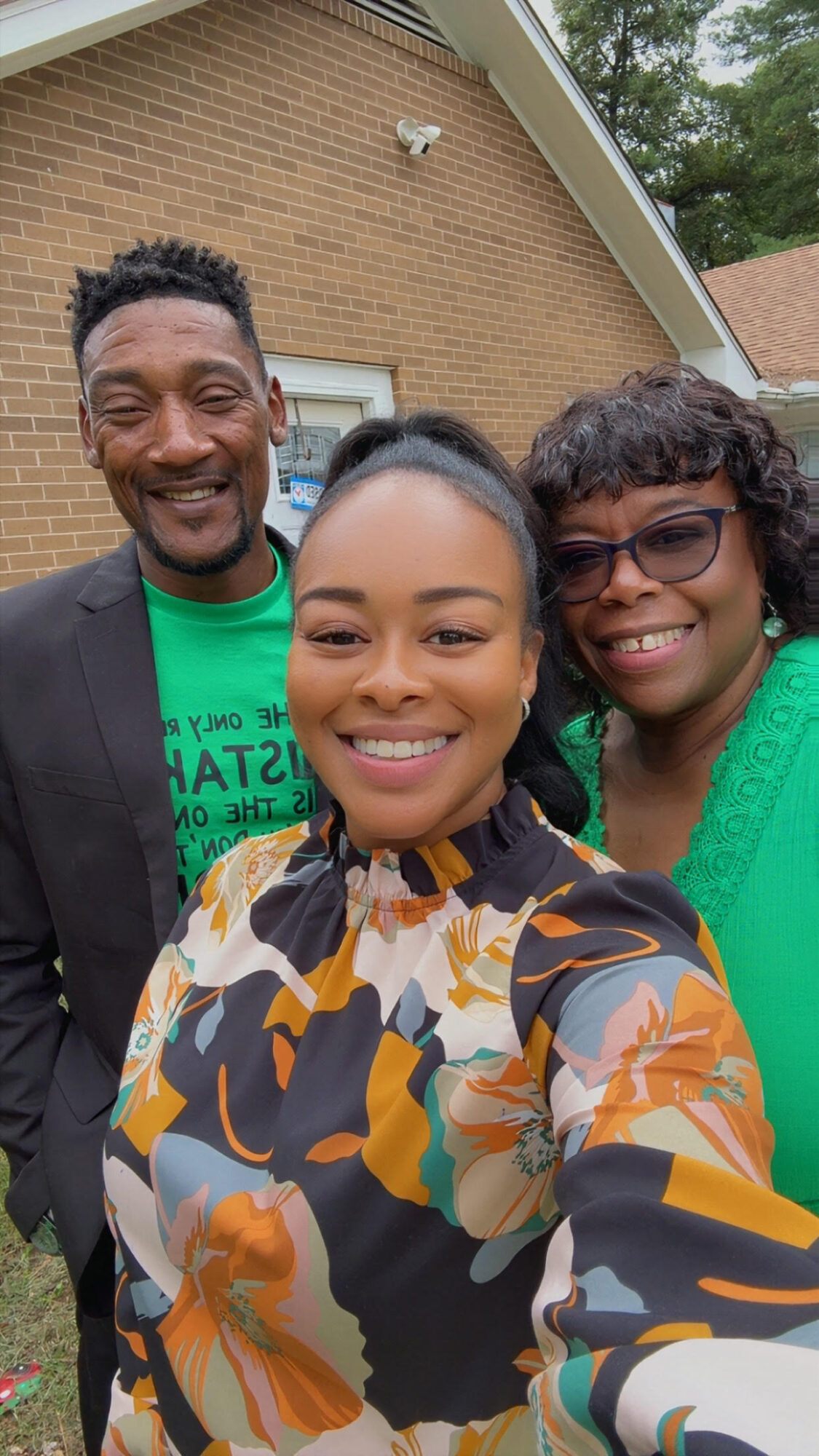 Three people smiling outdoors in front of a brick house, taking a selfie together.