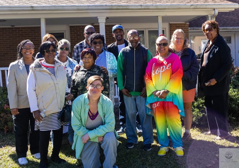 Group of diverse people standing outside in front of a house, smiling, with sunlight and grass visible.