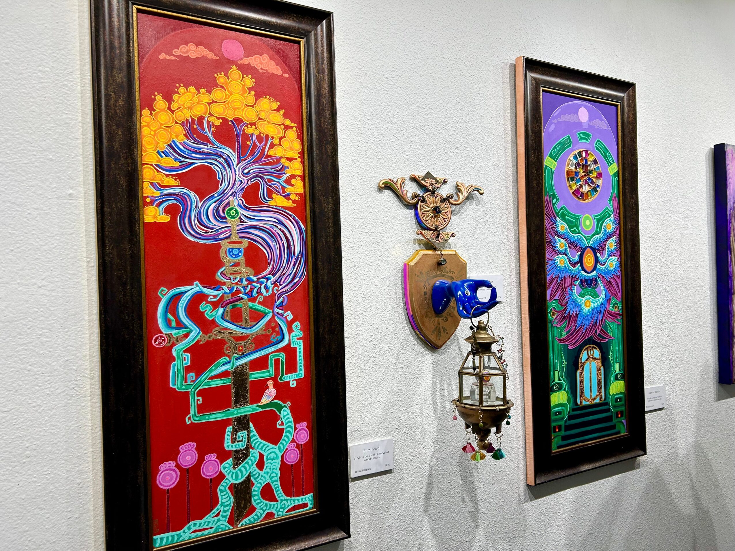 Two colorful framed artworks hang on a white wall, with a decorative wall-mounted sculpture between them, featuring a blue object and a small lantern.