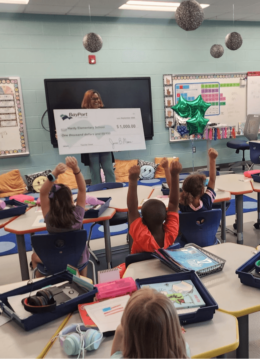 Classroom with students raising hands, teacher holding a large check, and a screen displaying the check. Students seated at desks with supplies.