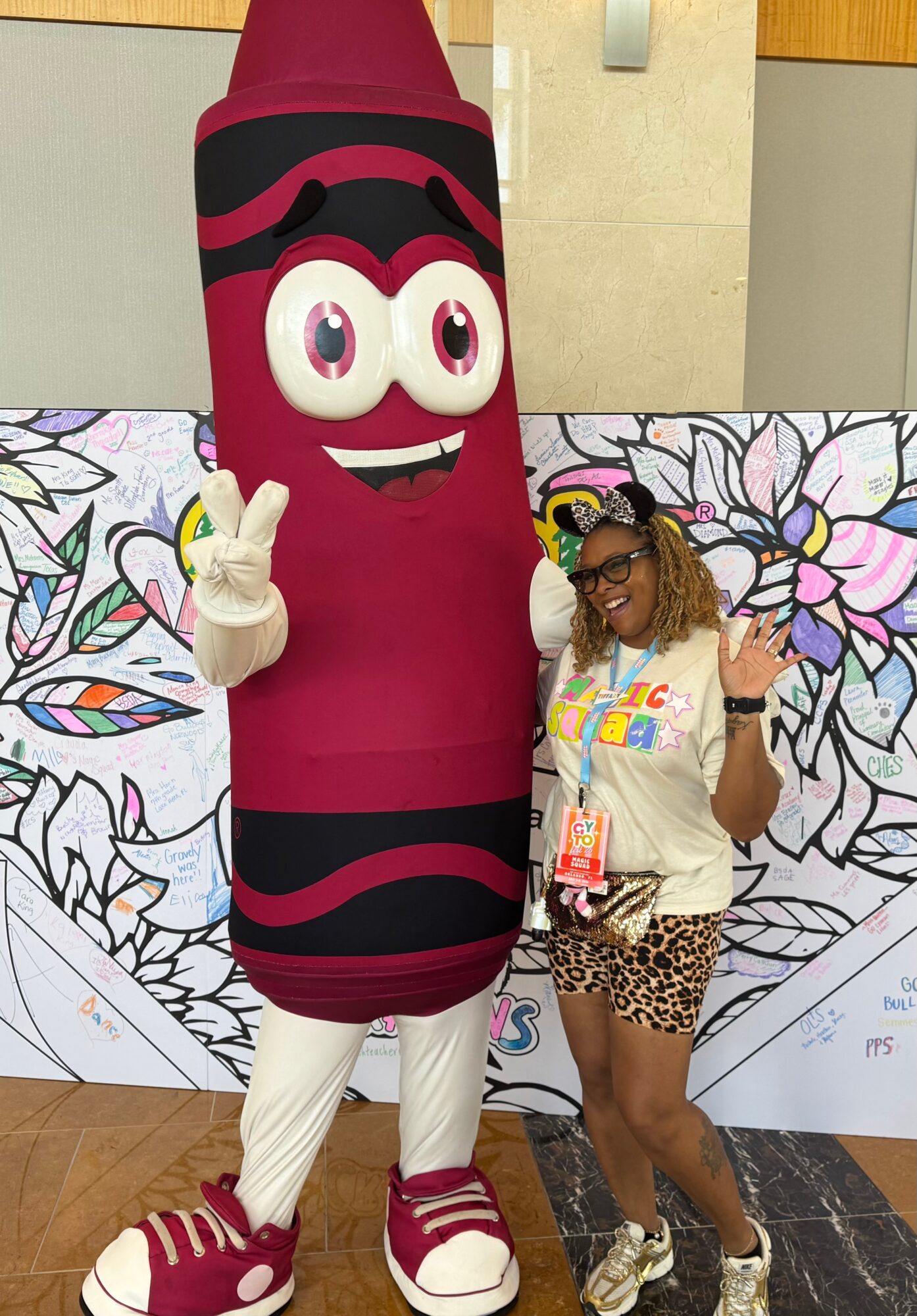 Person with curly hair and glasses standing next to a large cartoon crayon mascot, both making peace signs, in front of a colorful backdrop.
