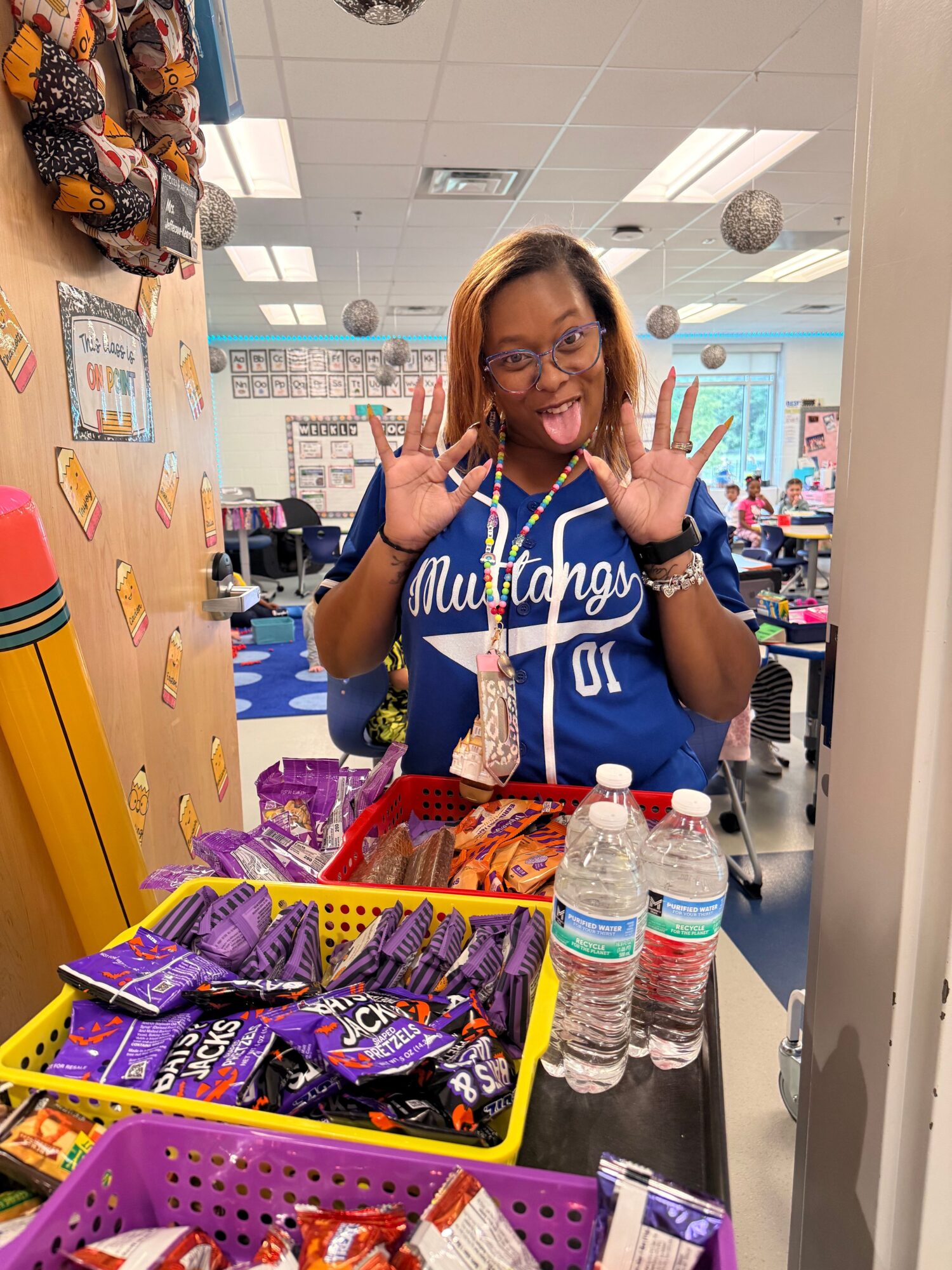 Woman in a blue shirt with white text, smiling and making peace signs, standing behind a table with snacks and water bottles, in a room with colorful decorations.