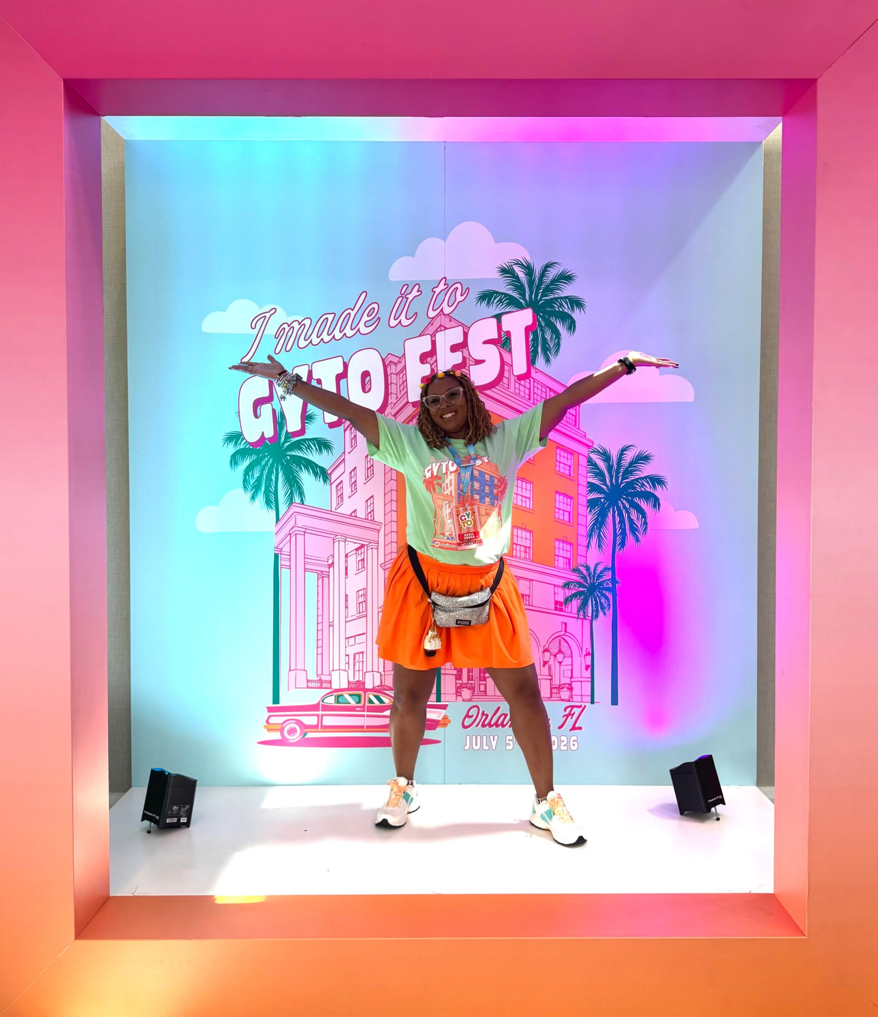 Person with arms outstretched standing on stage with colorful background and palm trees, smiling. Bright pink frame surrounds stage.