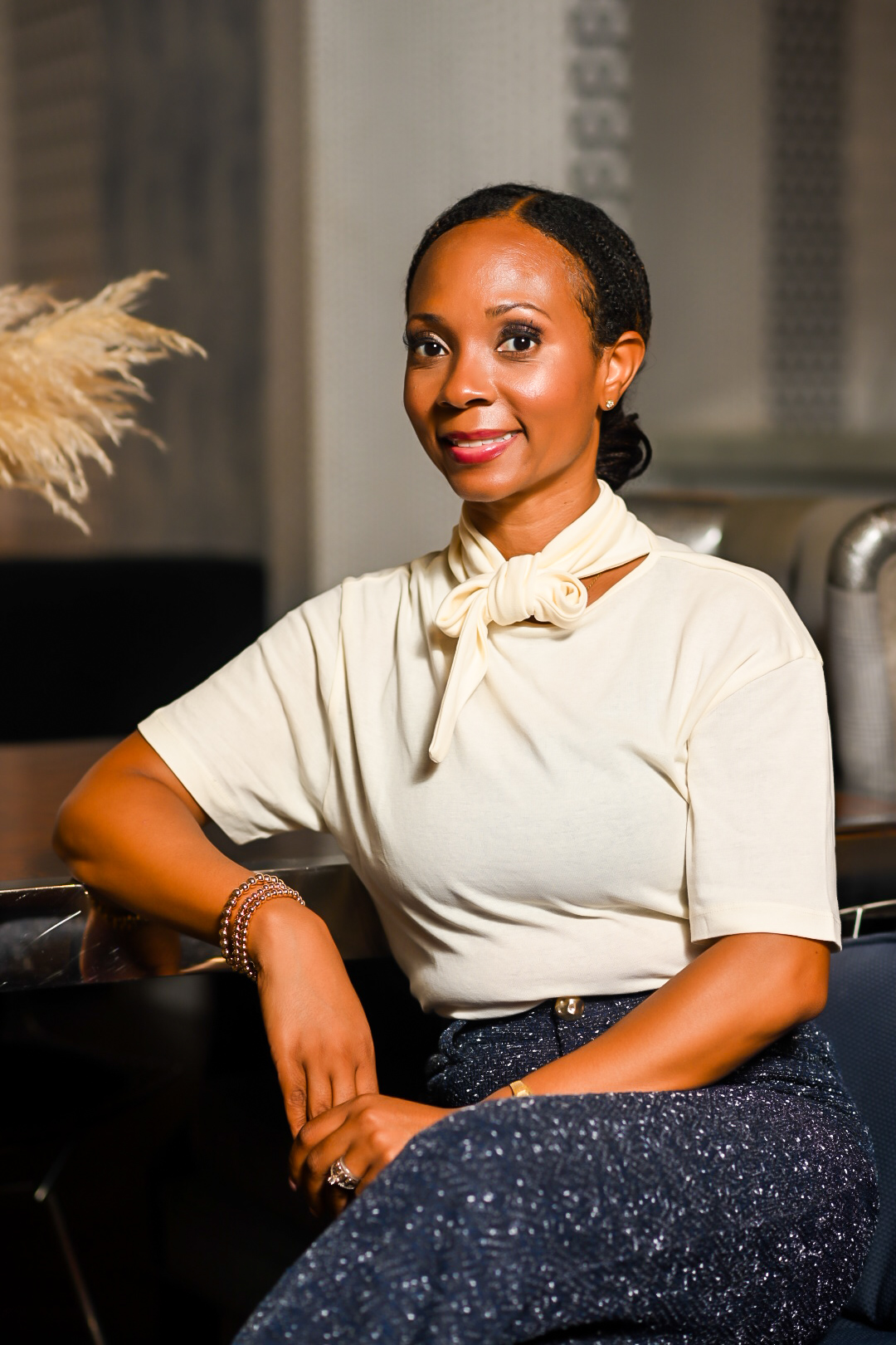 Woman with dark hair in a bun, wearing a cream blouse with a bow, sitting with her arm on a surface, smiling.