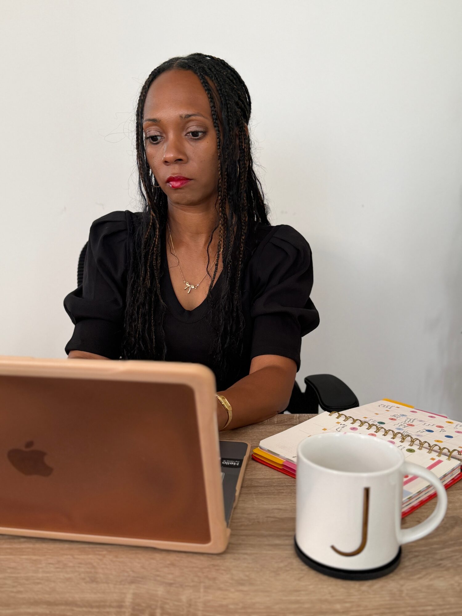 Woman with long dark hair sitting at a desk, looking at a laptop, with a mug and notebook nearby.