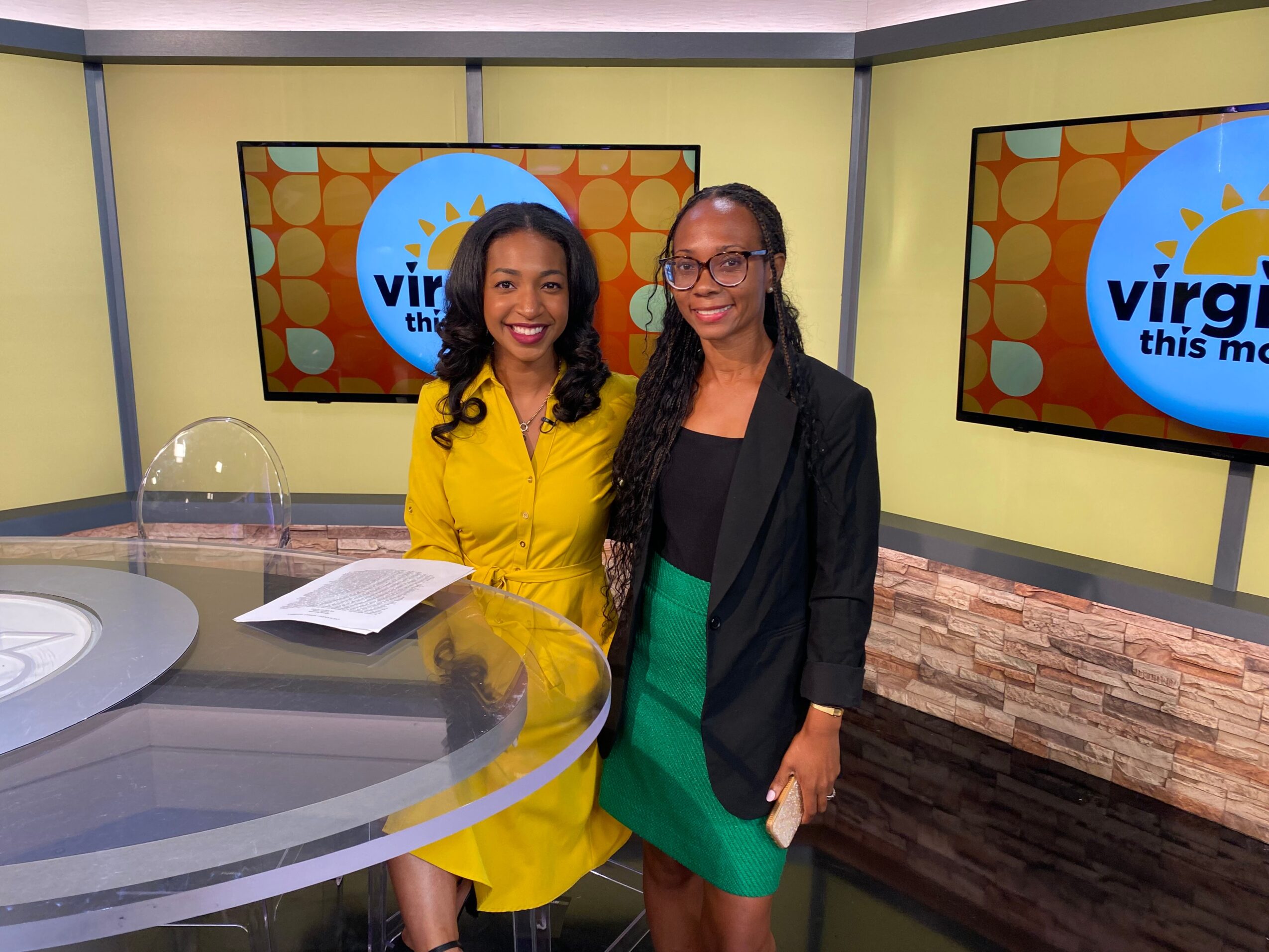 Two women standing together in a TV studio, smiling, with screens displaying a logo behind them.