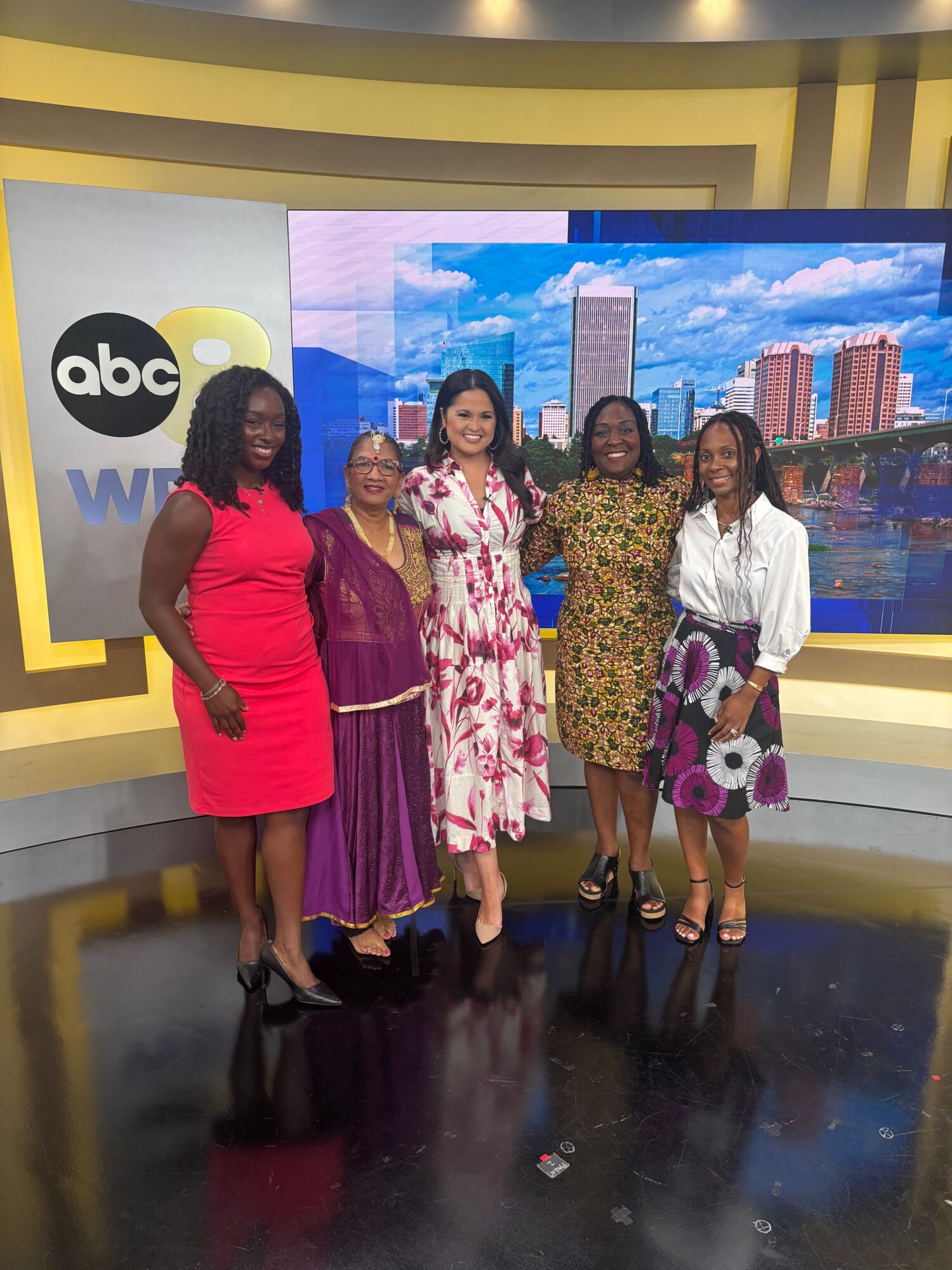 Group of six women standing in front of a large screen with ABC logo, smiling, dressed in colorful outfits.