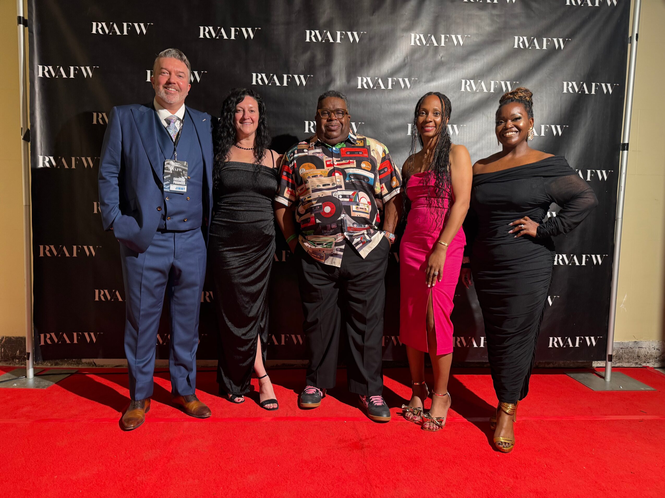 Five people standing on a red carpet in front of a black backdrop with white text, smiling and posing for the photo.