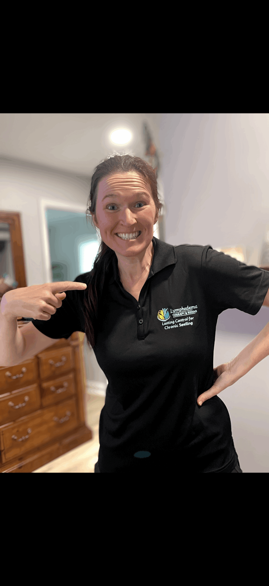 Smiling woman pointing at her black polo shirt with a logo, standing indoors with furniture in background.