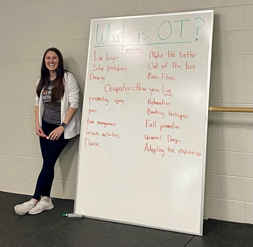 Young woman standing next to a whiteboard with notes about occupational therapy, smiling, in a hallway.