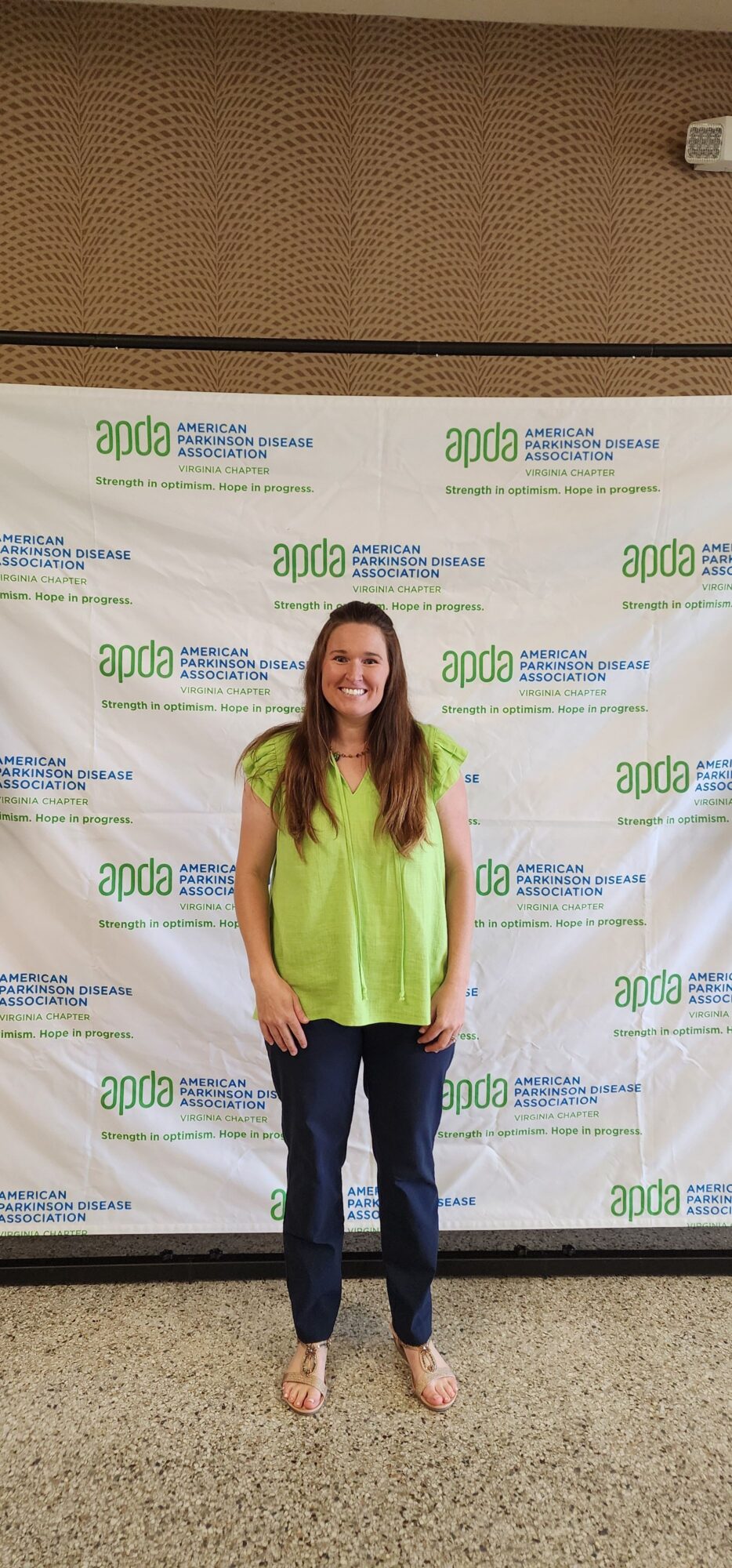 Woman standing in front of a backdrop with text, wearing a yellow top and dark jeans, smiling.
