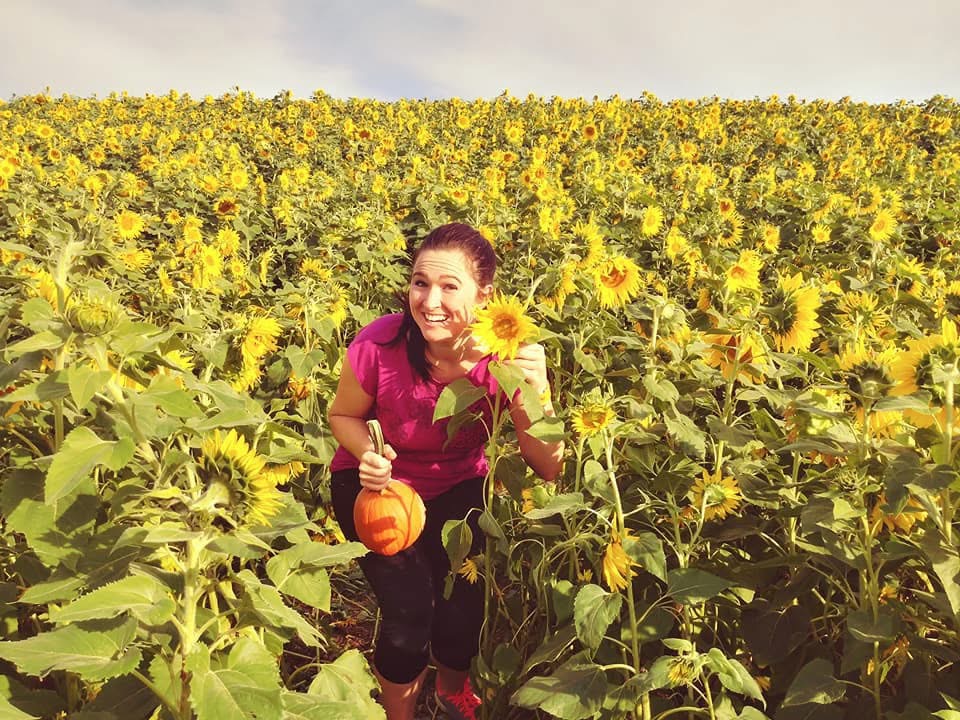 Woman smiling in a sunflower field holding a pumpkin, giving a thumbs-up, with a blue sky background.