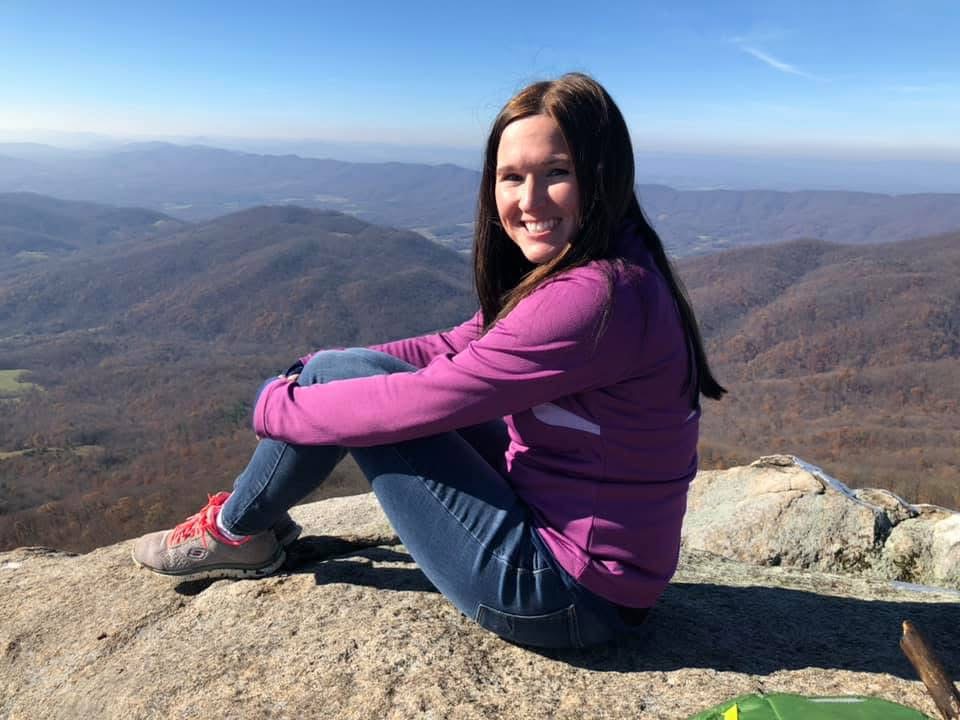 Woman sitting on a rock with mountains and sky in the background, smiling, wearing a purple jacket and jeans.