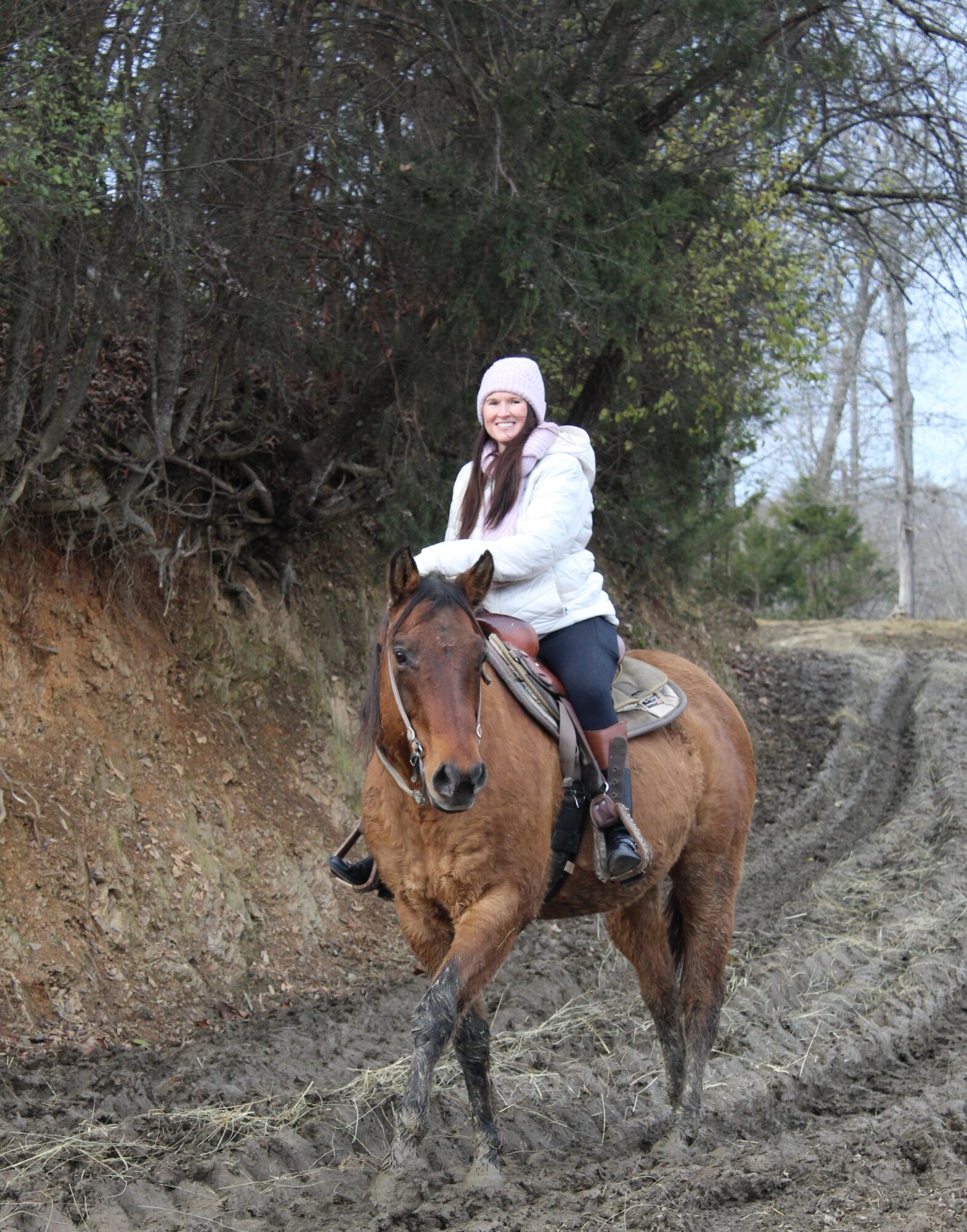 Person riding a brown horse on a dirt path with trees in the background, wearing a white jacket and hat.