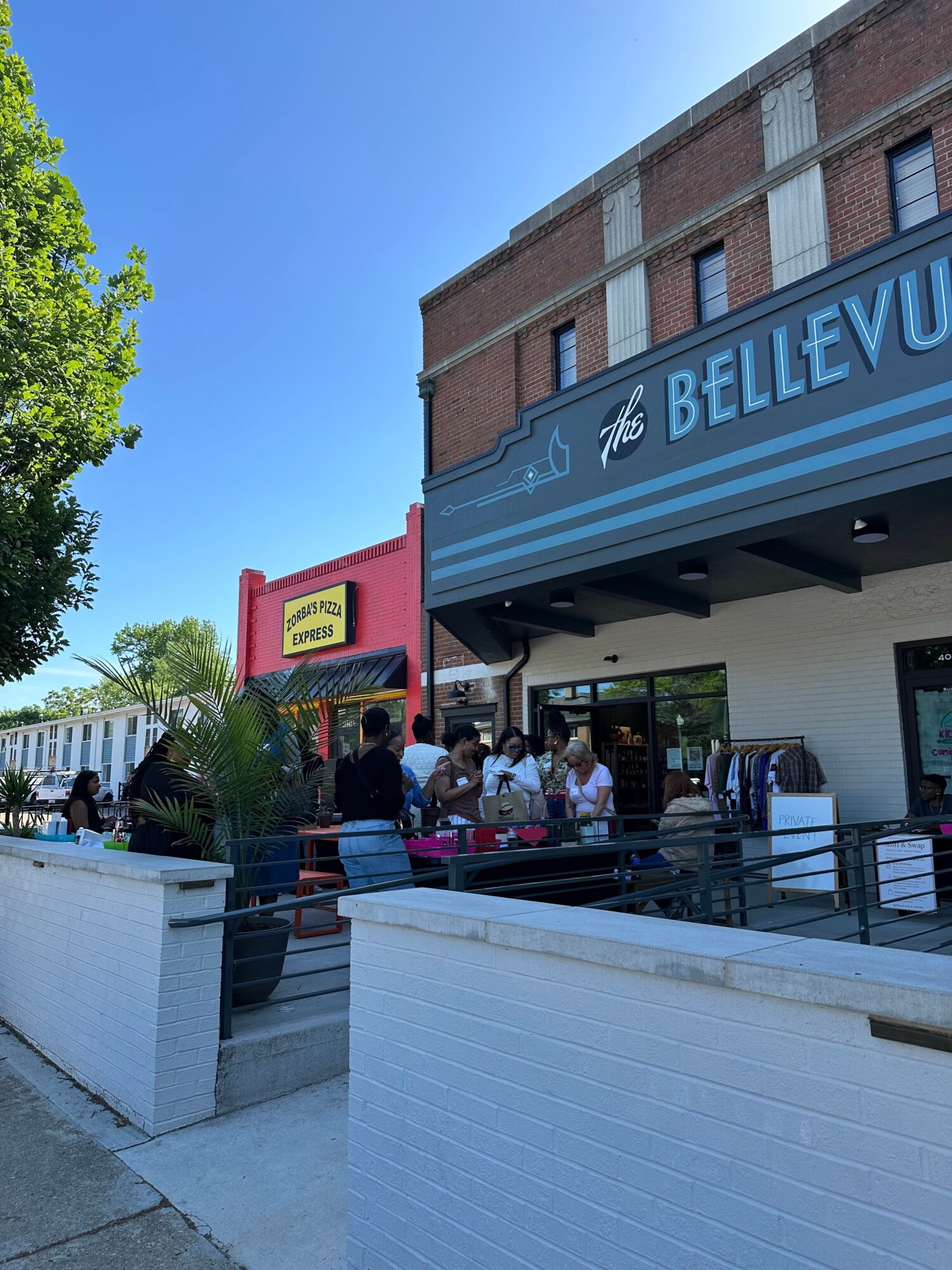People gather outside a building with a blue sign reading 'BELLEVUE' and a red sign, under a clear blue sky.