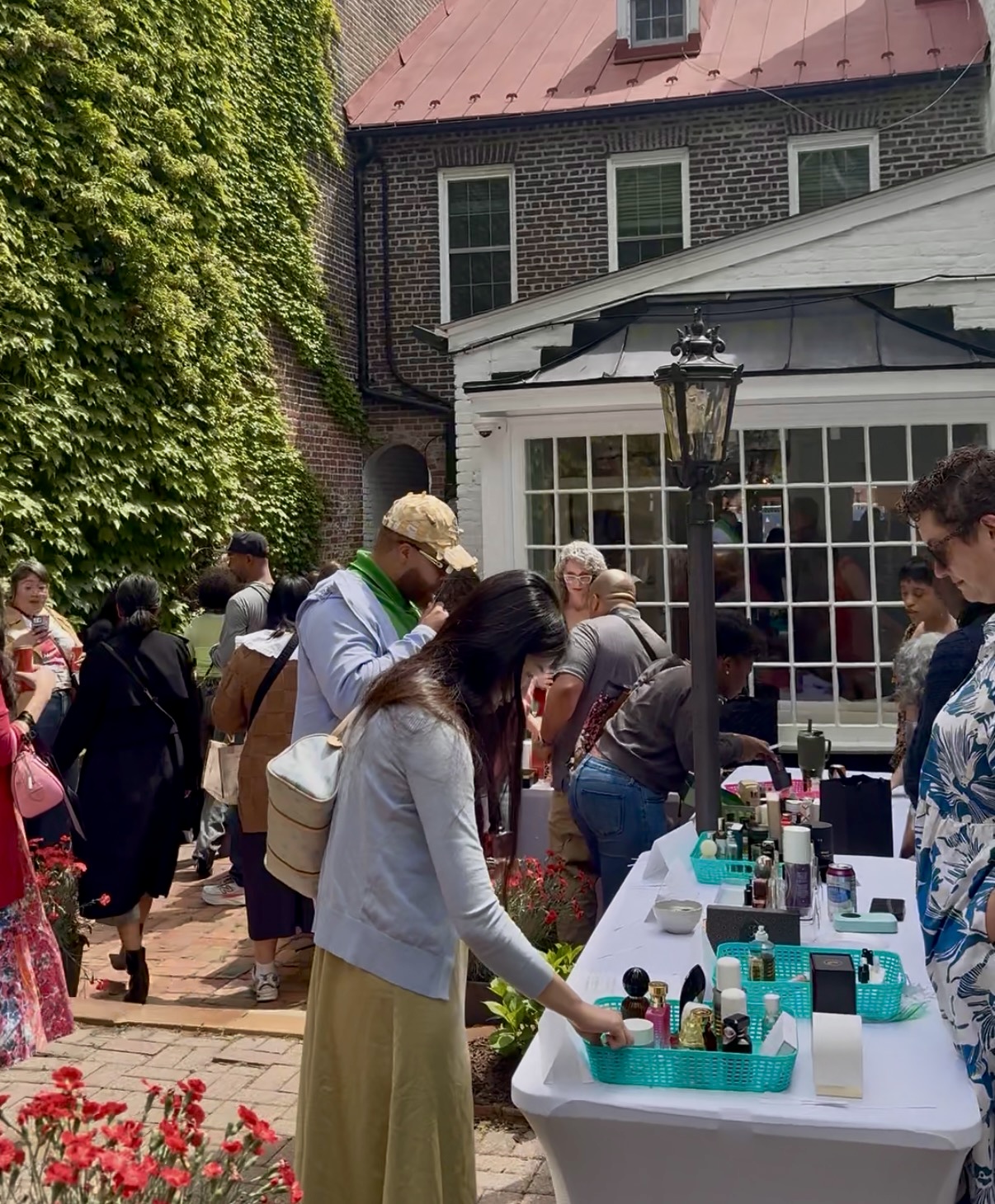 People gather outdoors around a table with bottles and items, near a building with large windows and a street lamp.