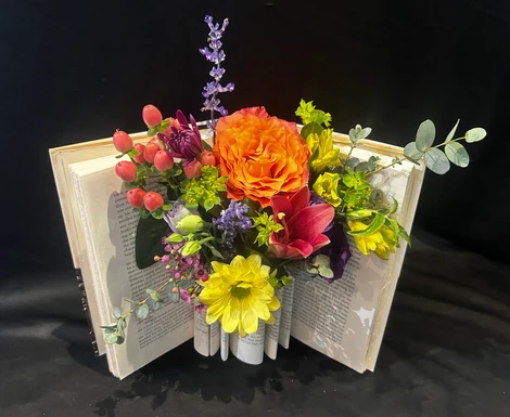 Open book with colorful flowers including orange, yellow, purple, and pink, arranged in the center, with green foliage and lavender accents.
