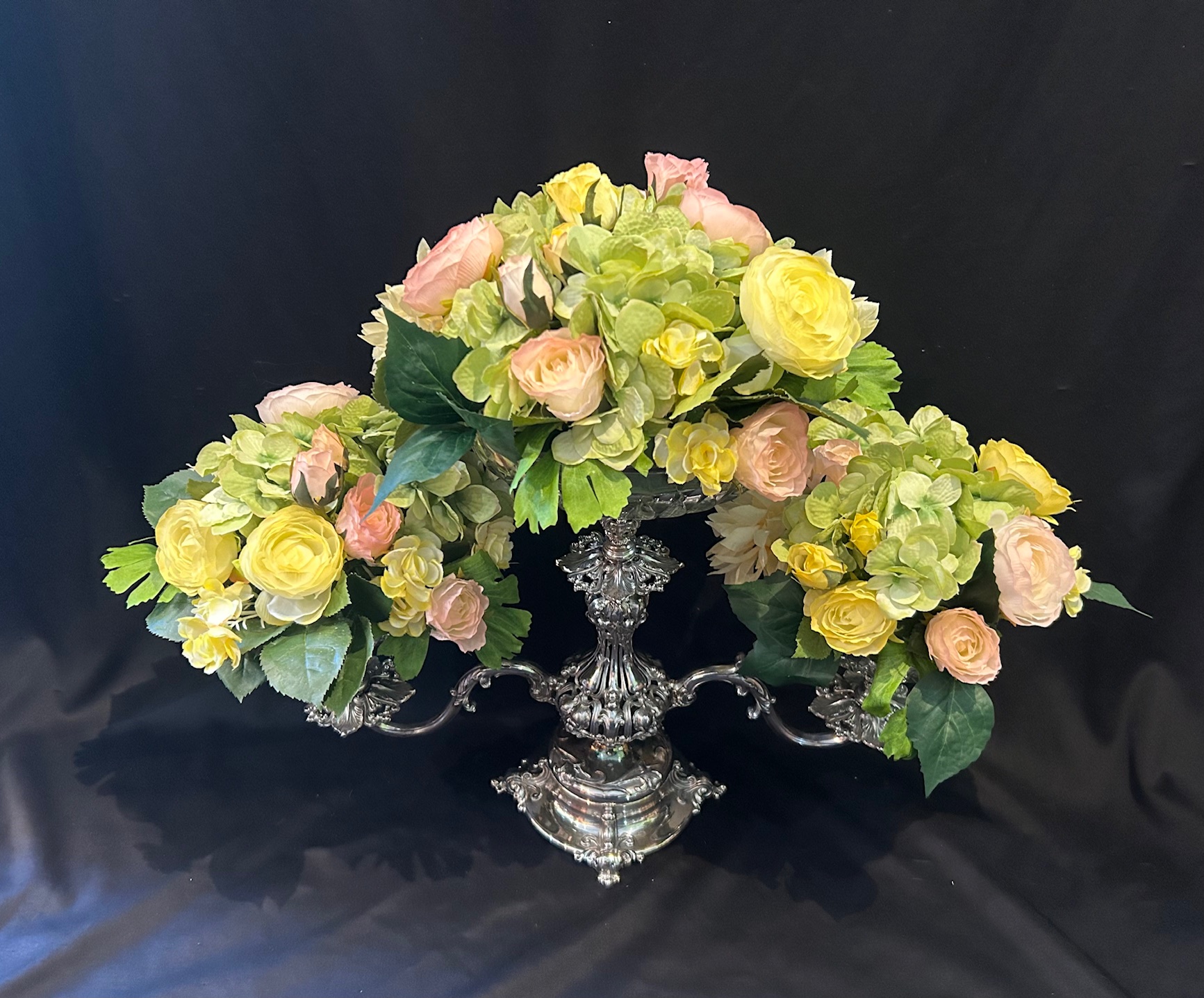 Silver ornate vase with pink and yellow flowers, green leaves, on black background.