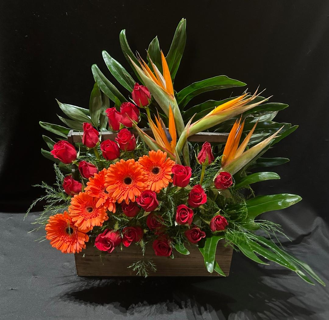 Arrangement of red roses, orange gerbera daisies, and bird of paradise flowers with green foliage in a rectangular container.