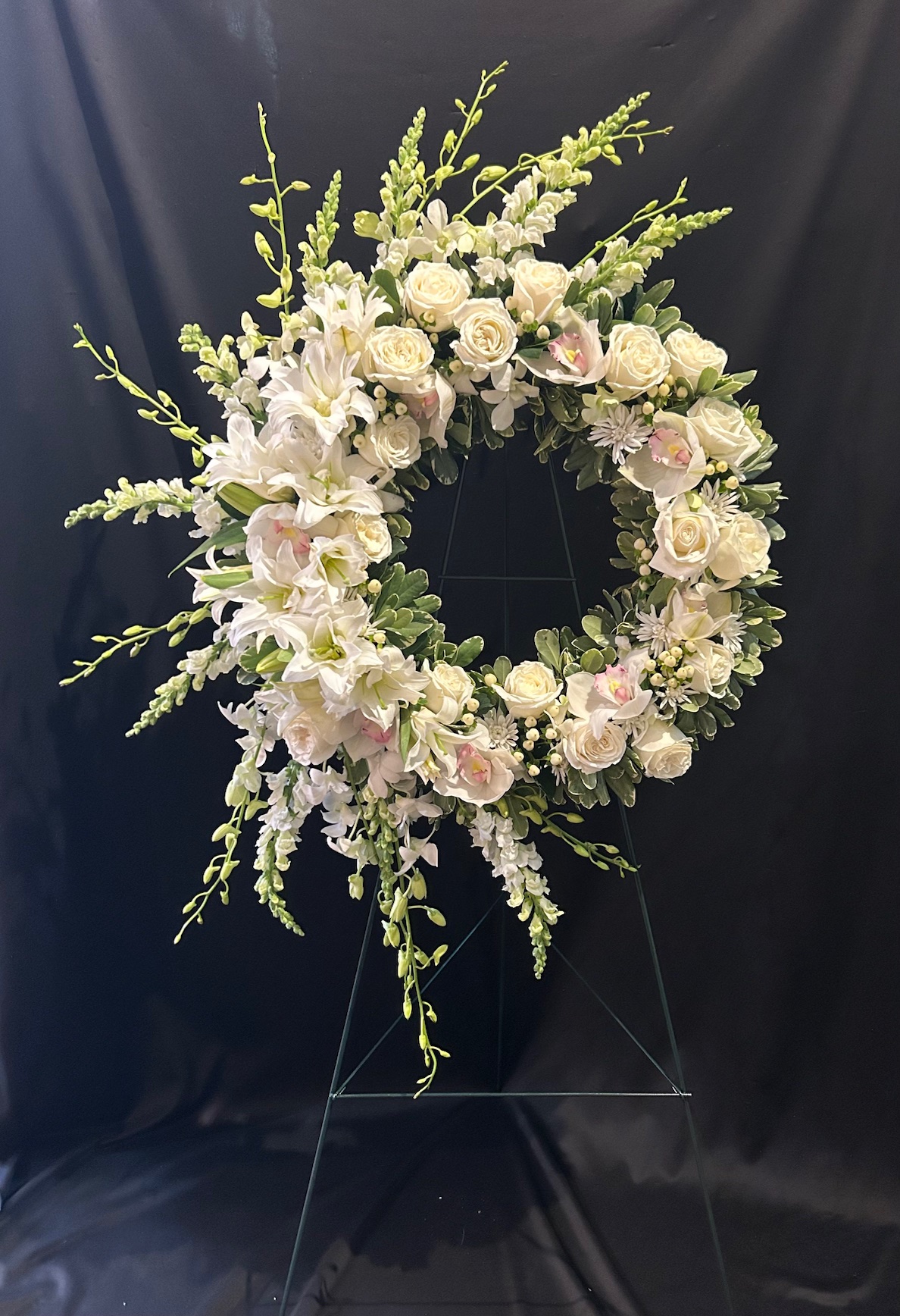 A floral wreath with white and pale pink flowers, green foliage, on a black background, displayed on a stand.