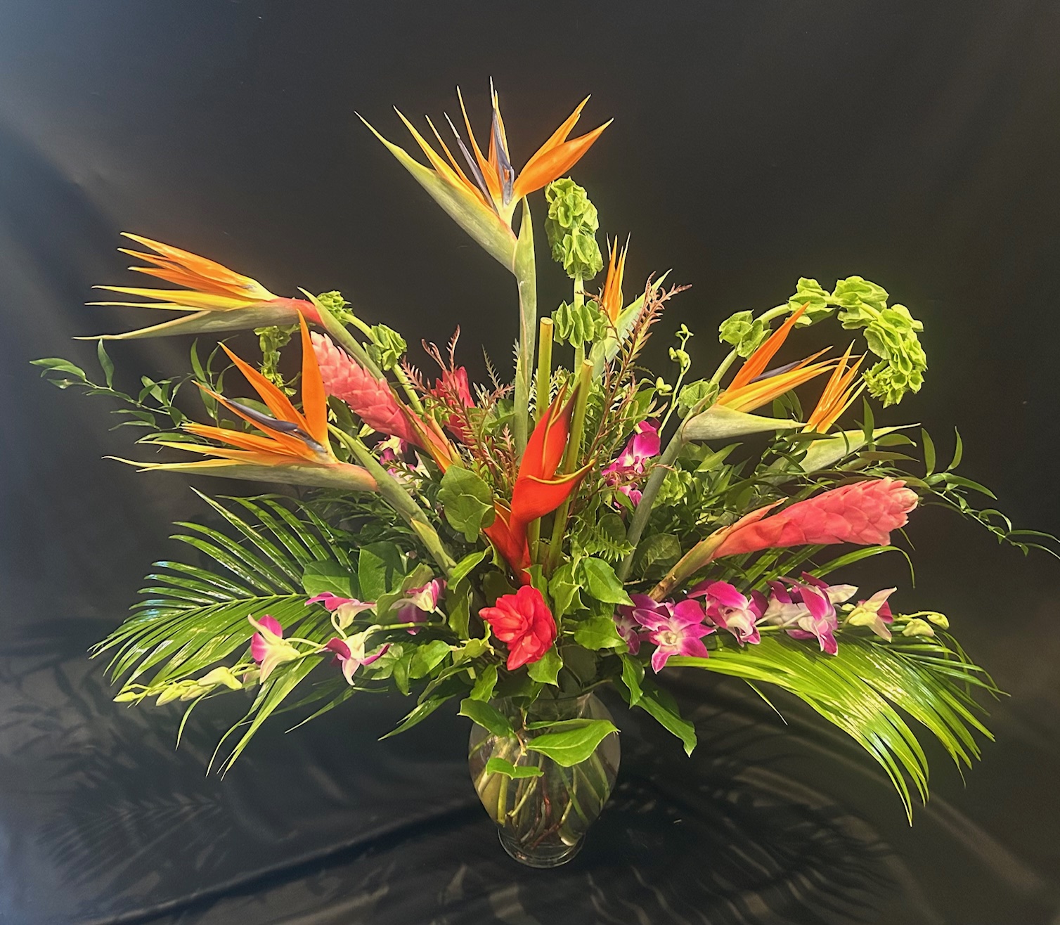 Colorful tropical flower arrangement with orange, pink, and green flowers and lush green leaves in a glass vase.
