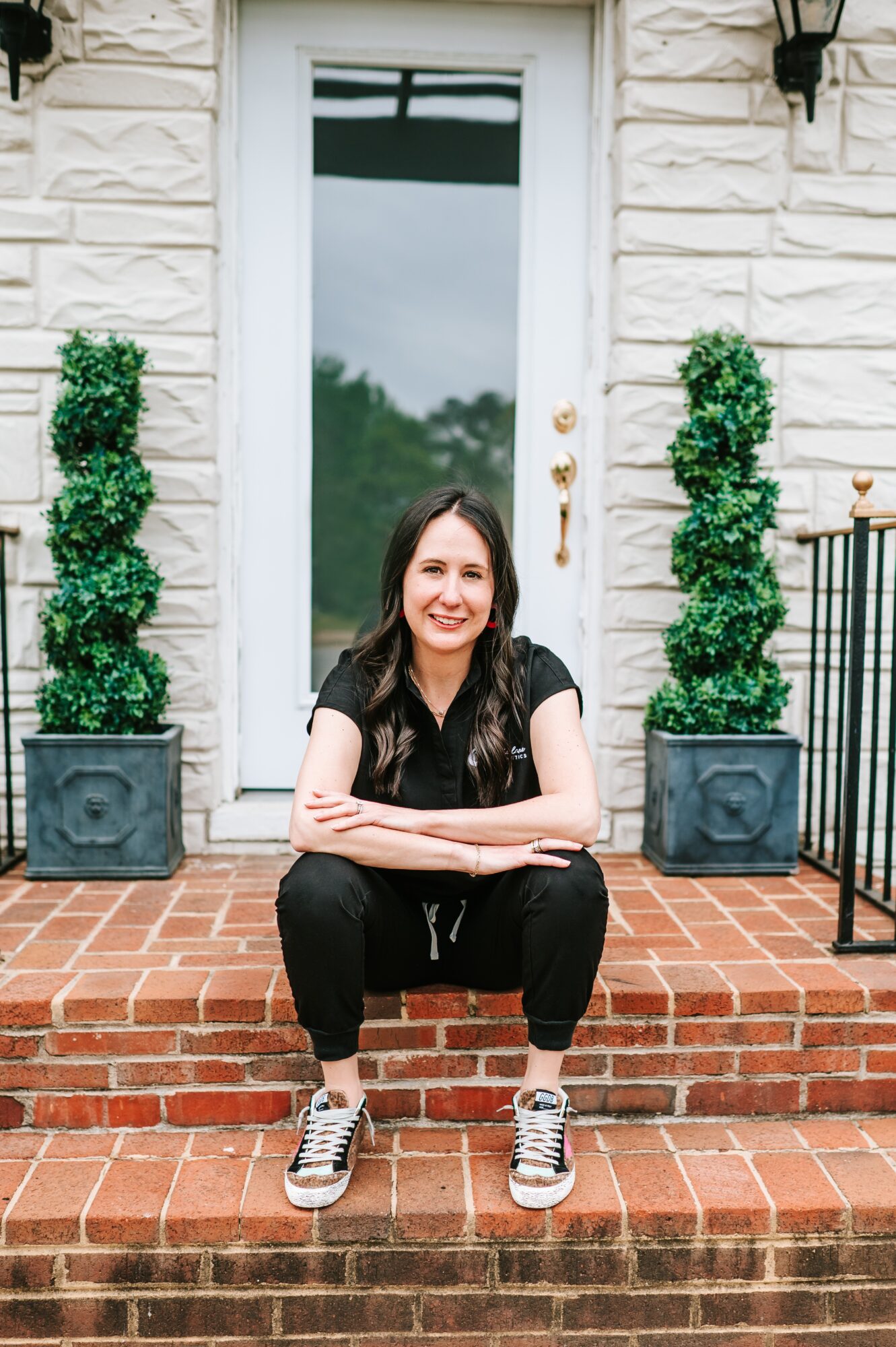 Woman squatting on brick steps in front of a door with two potted topiary plants on each side, smiling.
